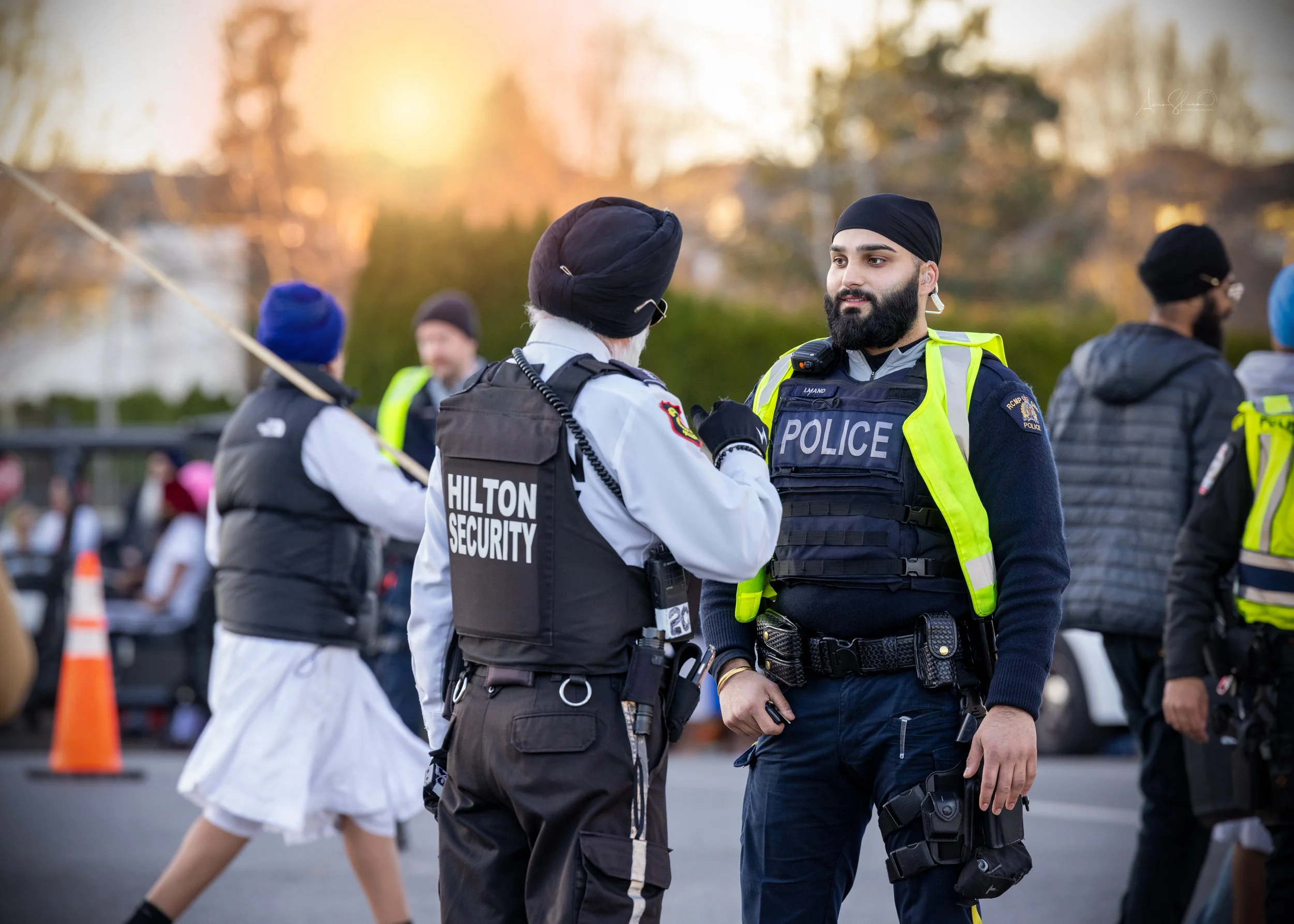Two police officers, one with a black turban and the other with a black vest labeled "Hilton Security," conversing during the daytime outdoor event, with other security personnel and people in the background.