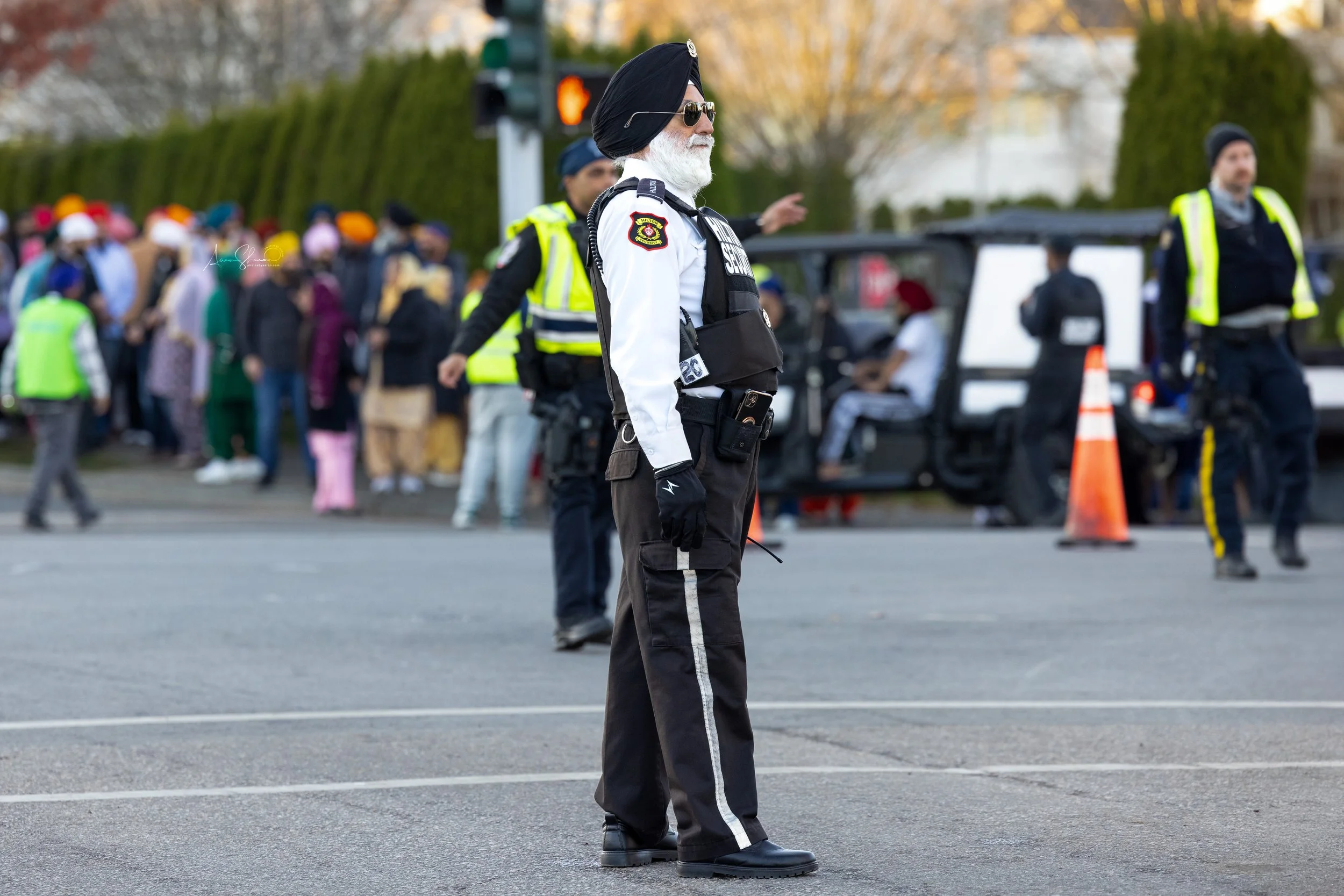 A police officer wearing a turban, sunglasses, and a uniform with a badge, standing in front of a crowd of people outdoors. The crowd appears to be gathering or waiting, with some individuals wearing colorful clothing and face masks. The scene is set