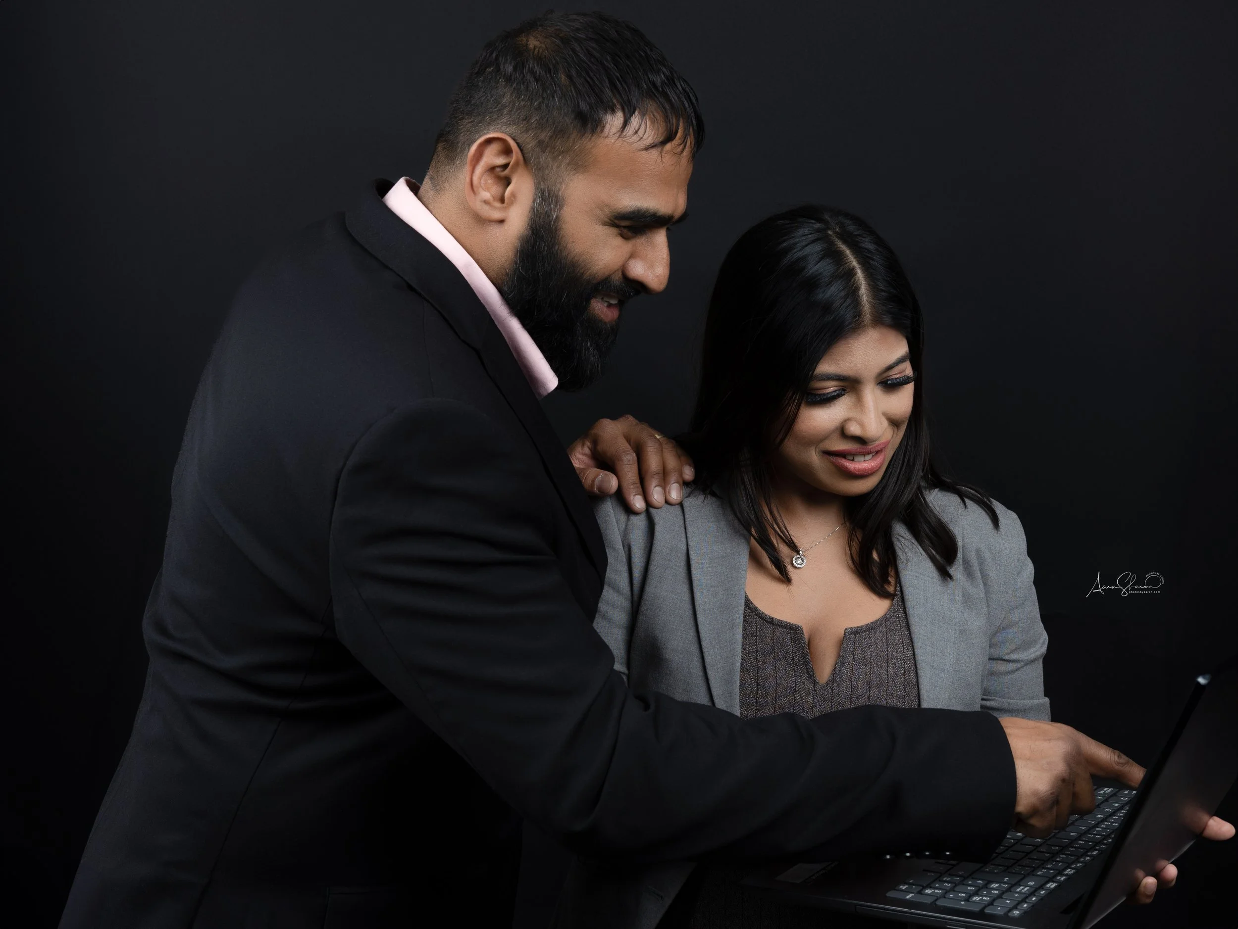 A man in a black suit sharing a moment of laughter with a woman in a gray blazer, as they look at a laptop together against a black background.