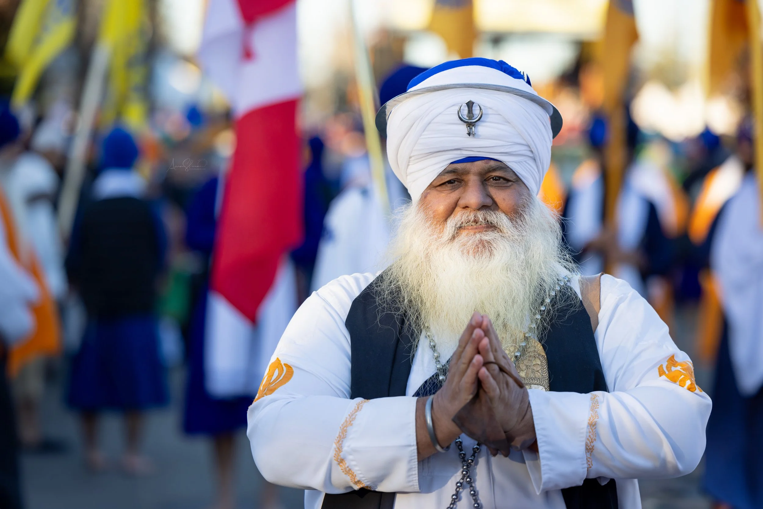 A man with a long white beard and traditional Sikh turban making a prayer gesture at a cultural parade, with other participants in the background holding flags.