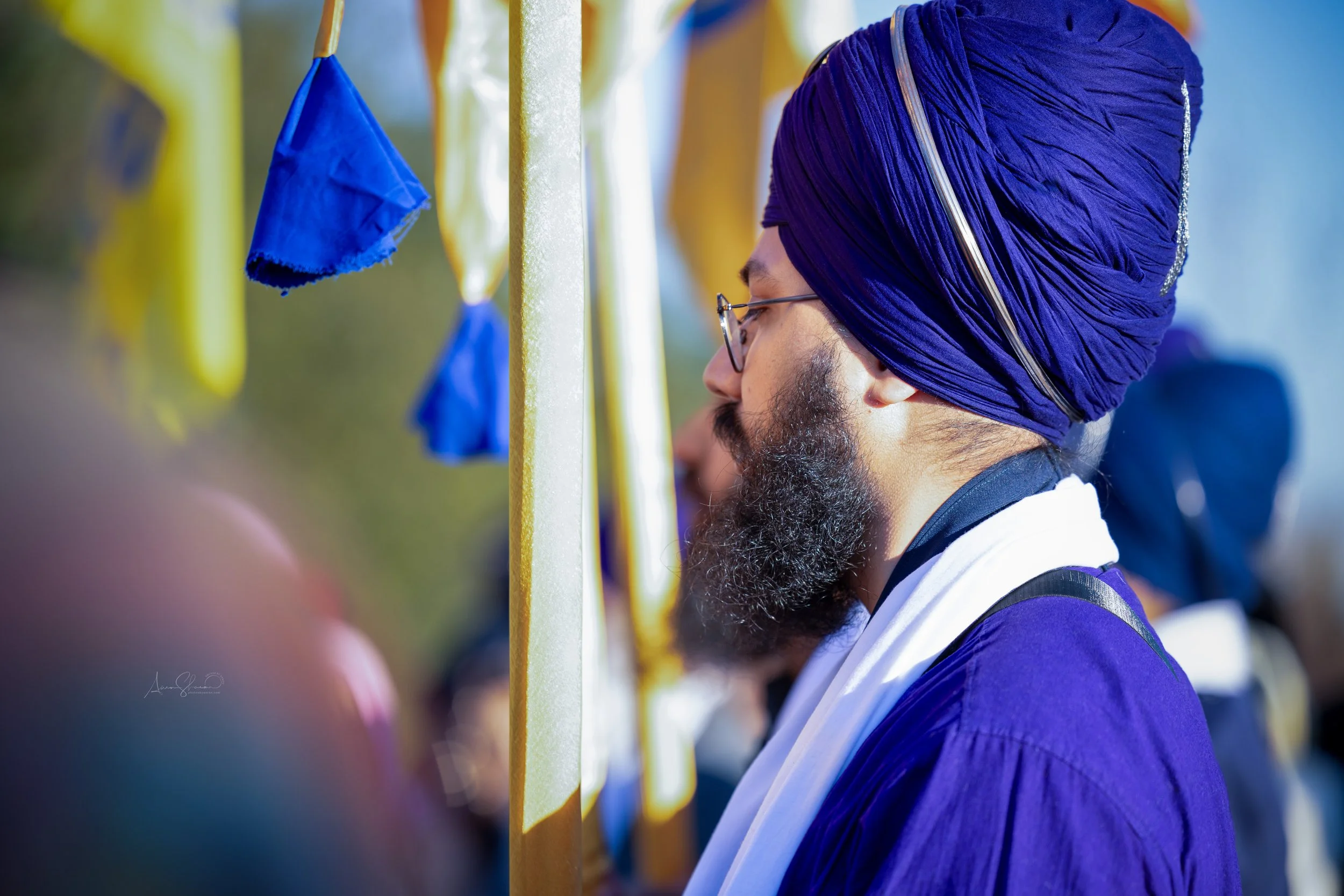A man wearing a purple turban, glasses, and a purple academic gown with a white stole, standing among other graduates at a graduation ceremony.
