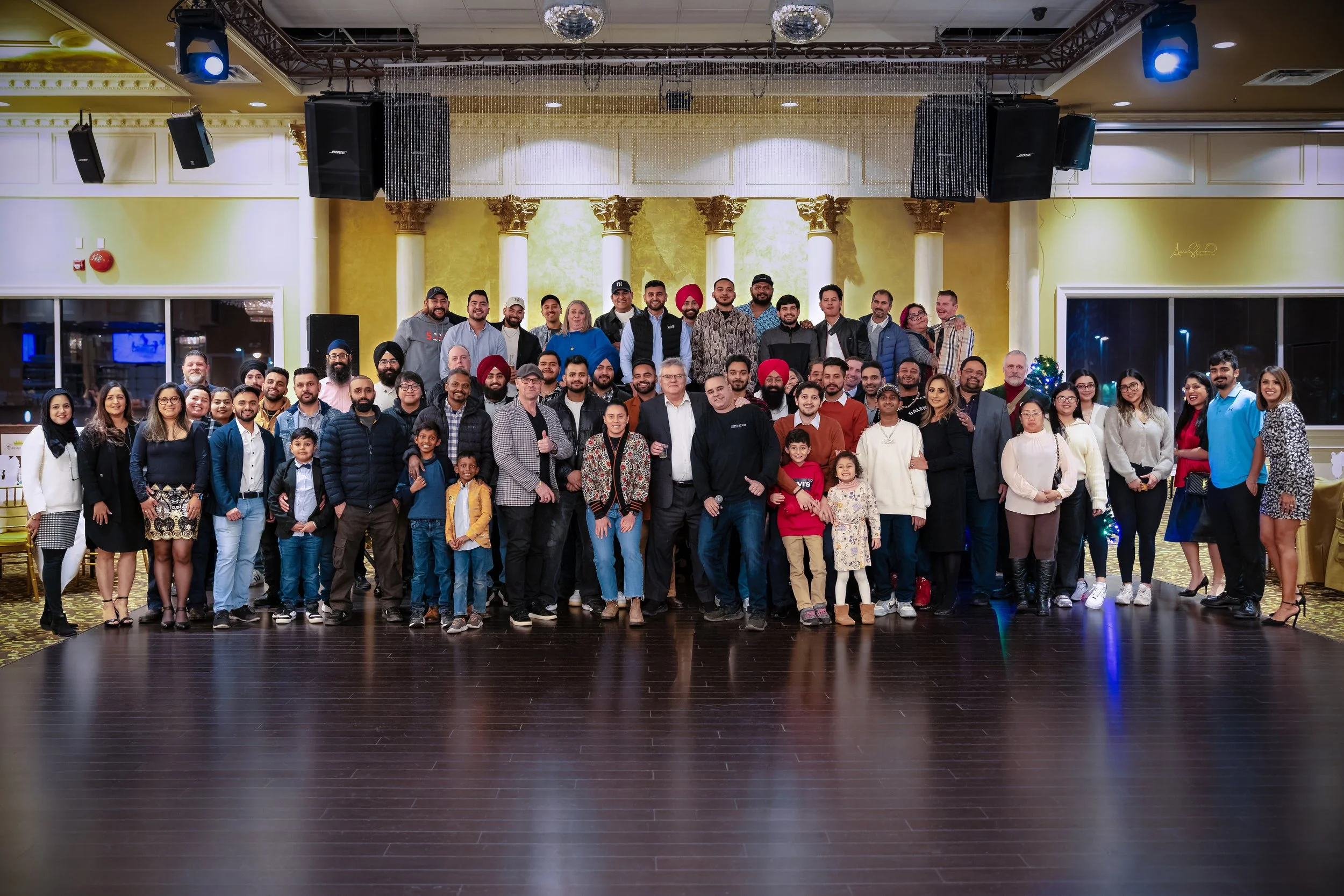 A large group of people gathered for a group photo in a decorated banquet hall with columns and a stage, including children and adults, some dressed casually, others in semi-formal attire.
