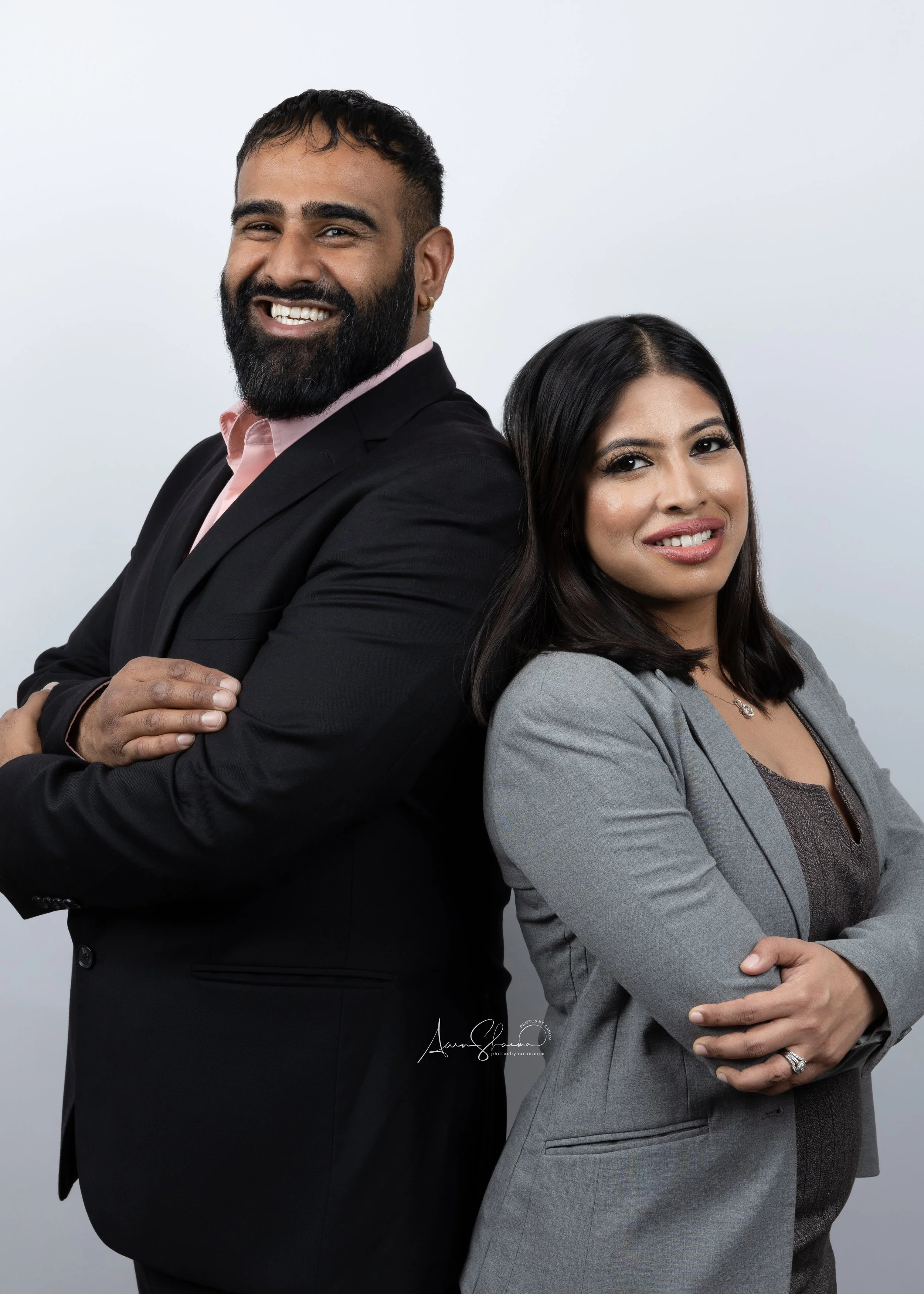 A man and woman standing back-to-back with arms crossed, smiling confidently, against a plain light gray background.