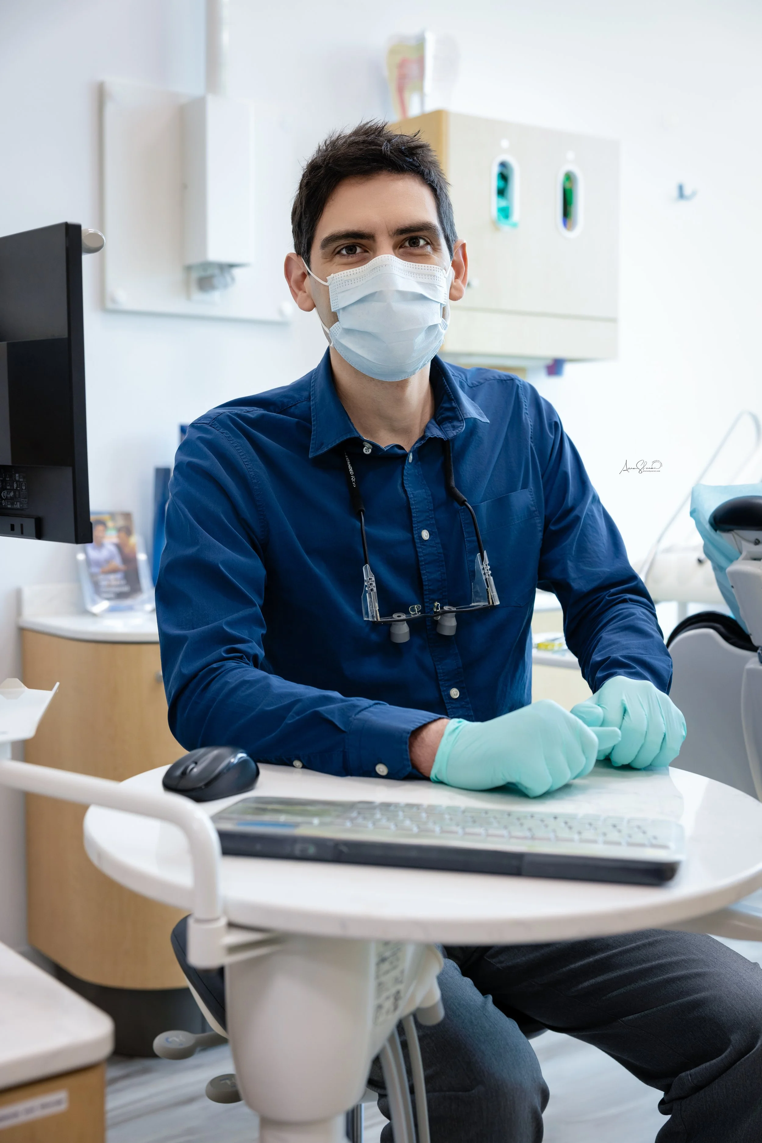 A male dentist wearing a face mask, gloves, and glasses around his neck, sitting at a dental clinic, smiling and looking at the camera.