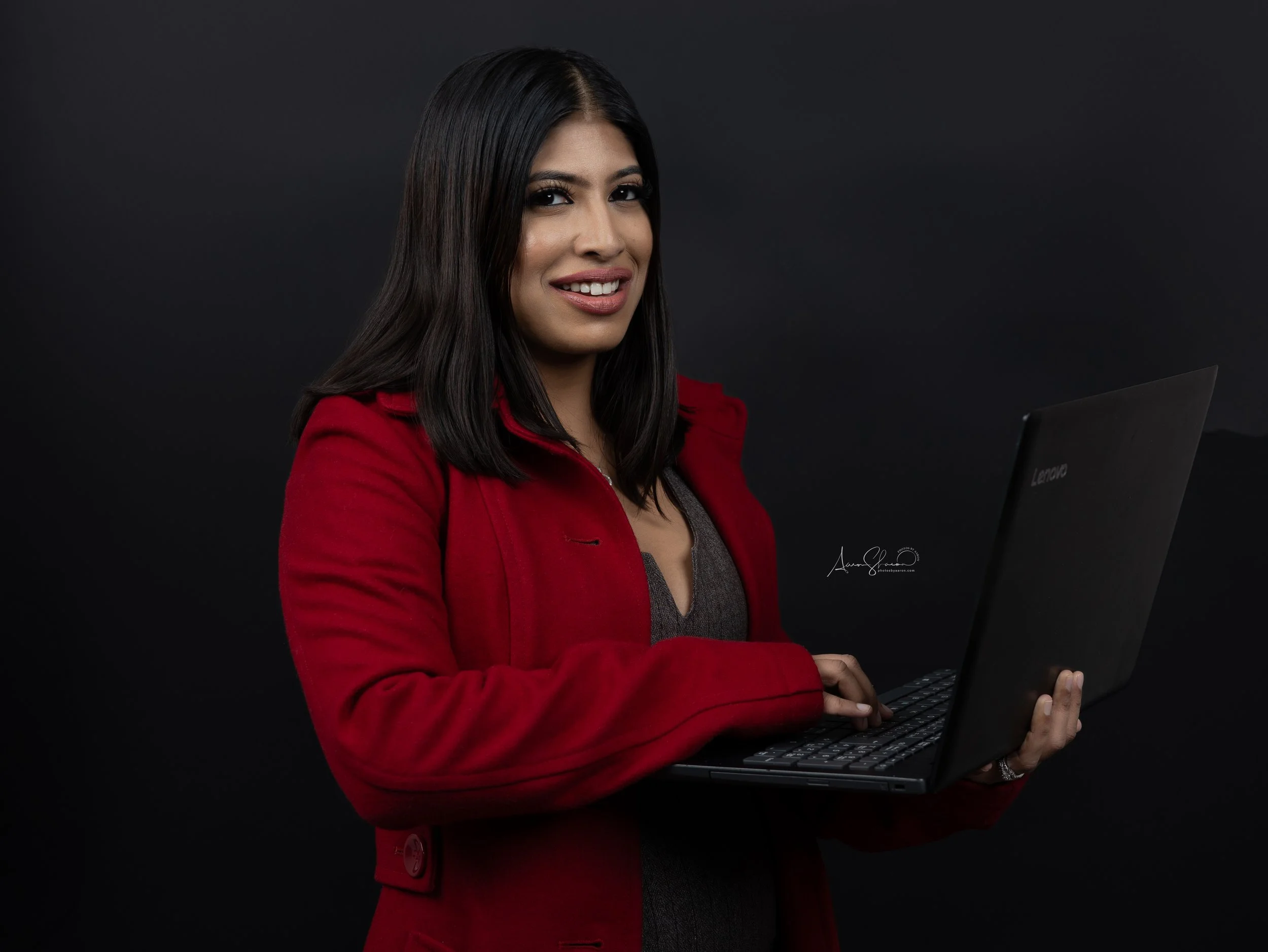 A woman with long dark hair wearing a red blazer and gray top holds a black laptop against a dark background.