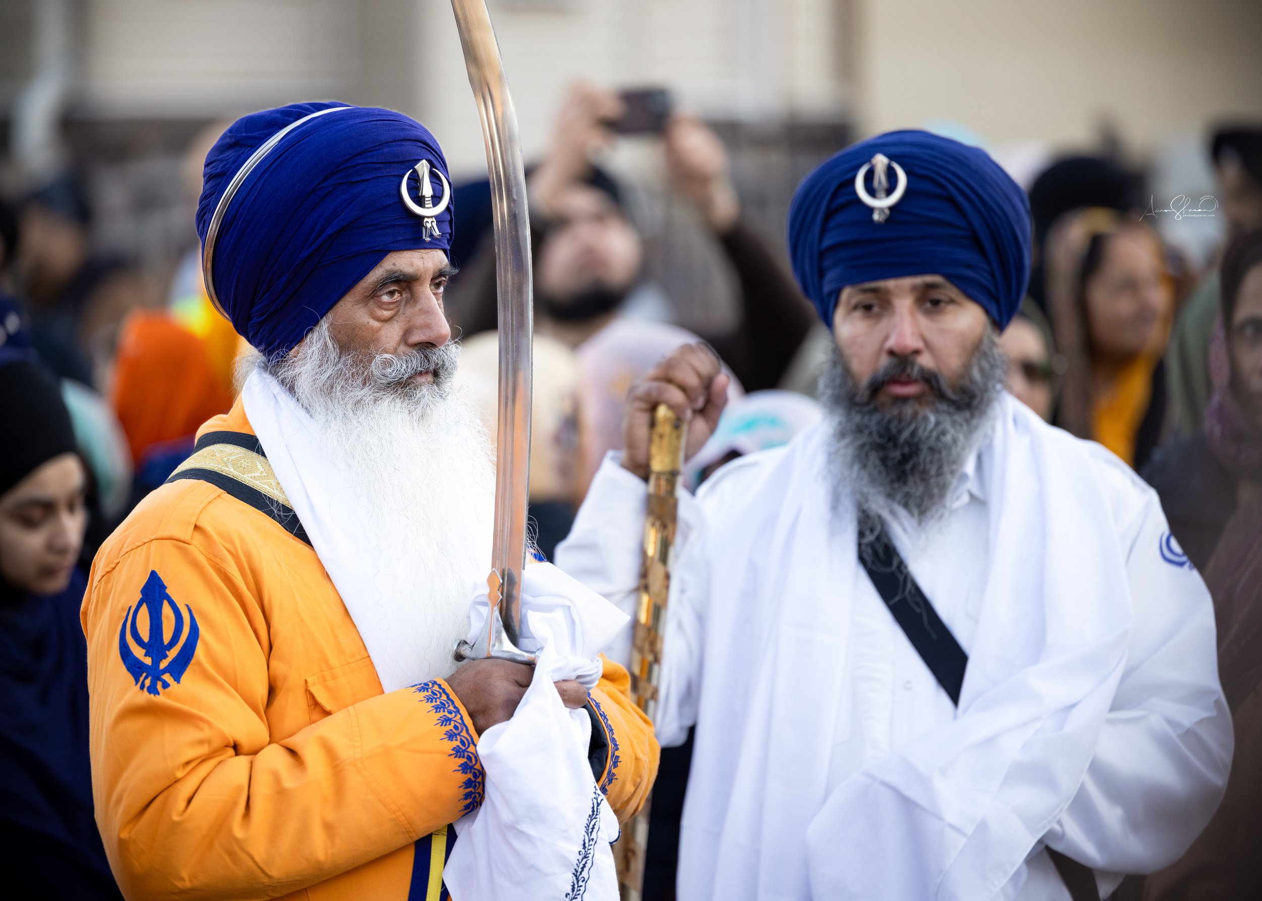 Two Sikh men wearing blue turbans and white attire, holding swords, surrounded by a crowd of people.