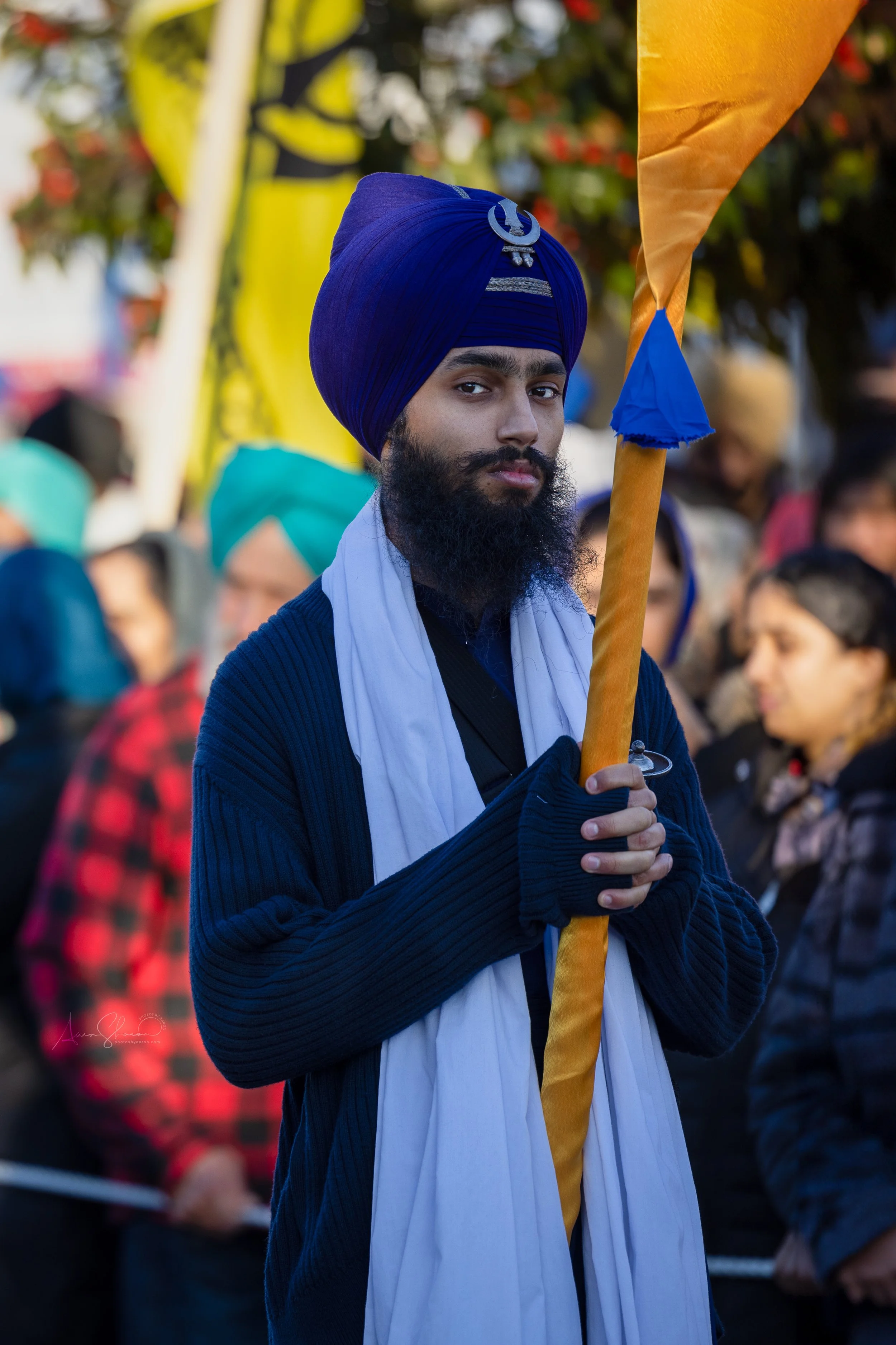 A man wearing a blue turban and a white scarf holds a saffron-colored flagpole during a gathering or protest, with other people in the background.
