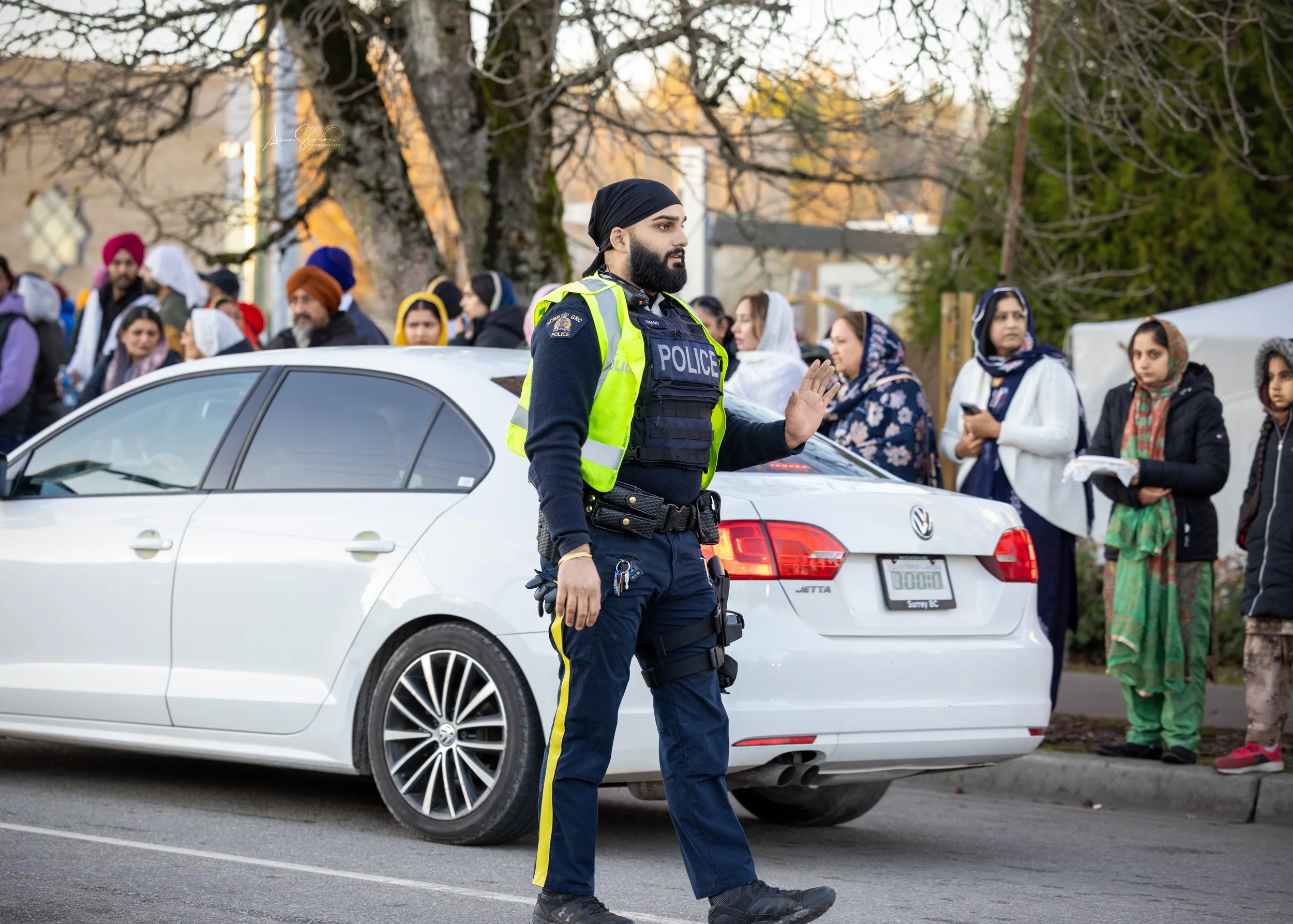 A police officer standing in front of a white Volkswagen Jetta, gesturing with his hand, while a group of diverse people, mostly women, stand behind him on the sidewalk.
