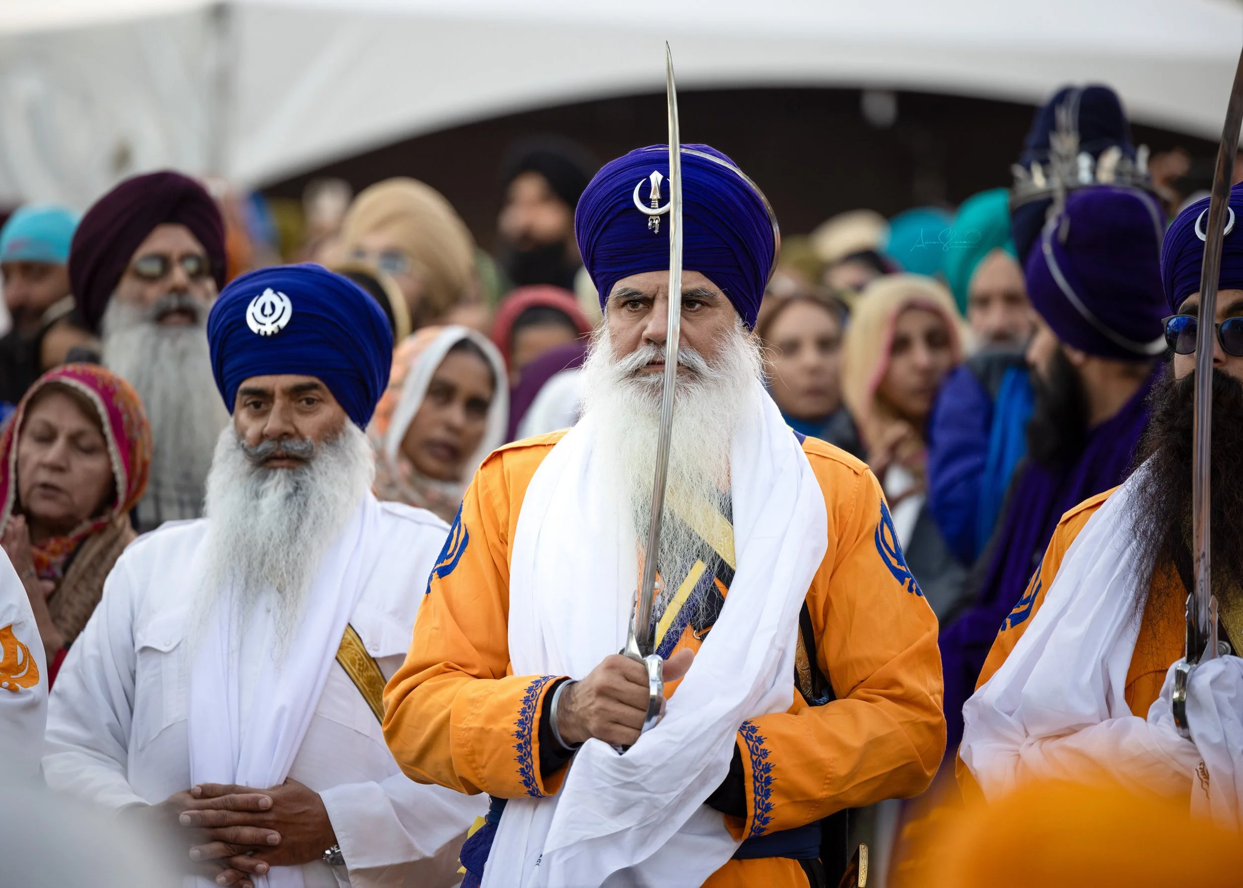 A man with a long white beard wearing a blue turban and orange traditional attire holding a sword during a gathering of people, many also in colorful turbans and traditional clothing, in an outdoor setting.