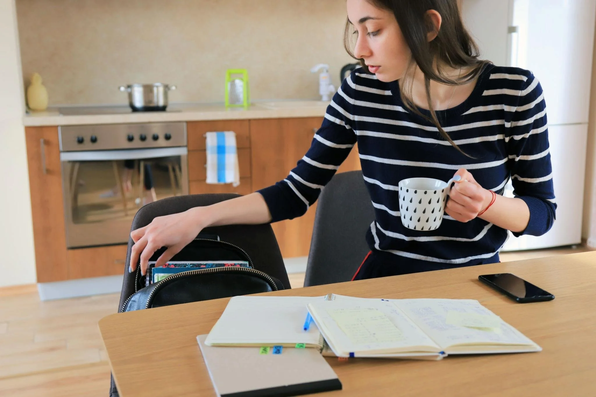 Person packing backpack at a kitchen table while holding a mug, with notebooks and a planner open during morning routine