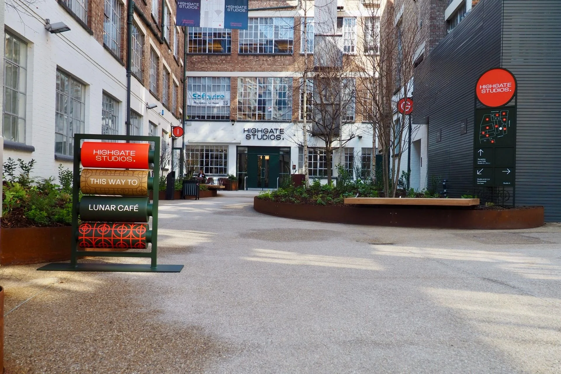 An outdoor courtyard area of Highgate Studios with street signs and pathways, surrounded by brick and modern buildings, trees, and benches.