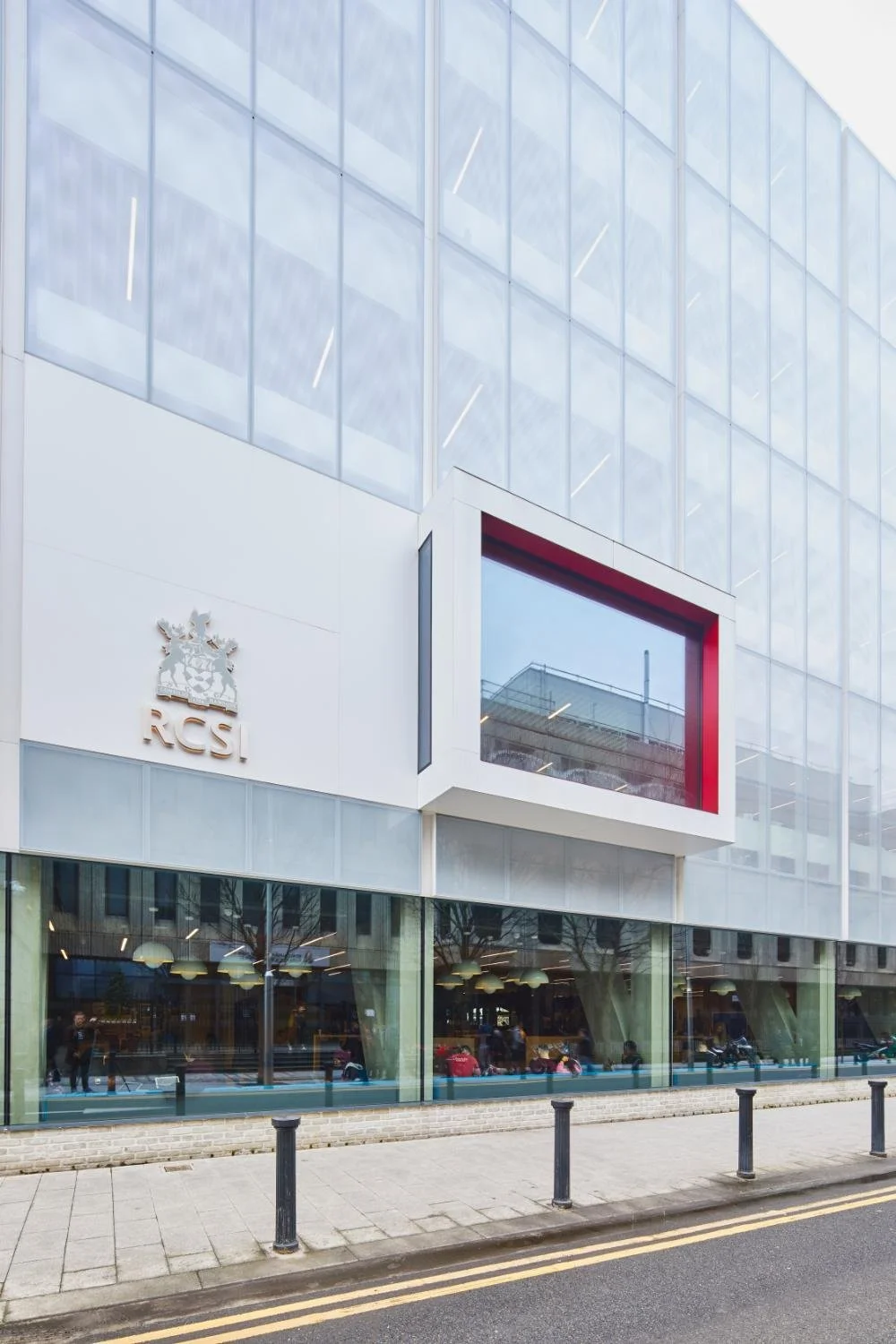 Modern building with large glass windows and a white facade, displaying the RCSI logo and crest. The street in front has bollards and a sidewalk.
