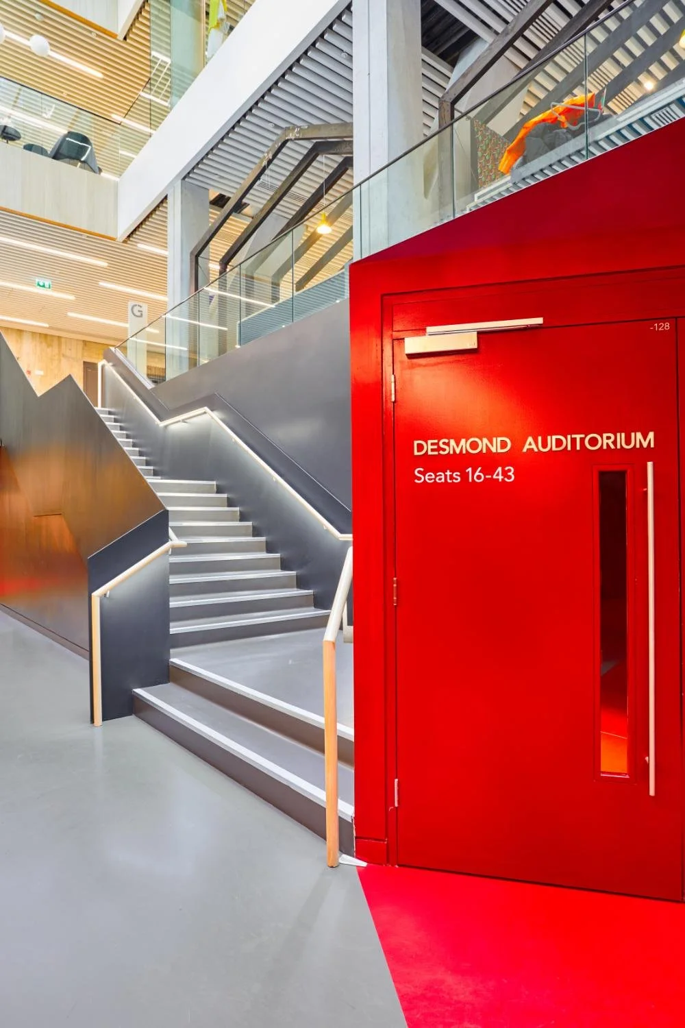 Modern indoor staircase with gray steps and white handrails next to a bright red door labeled 'Desmond Auditorium' with seating for 16-43. The space features glass railings, mixed wood and metal finishes, and an open, contemporary design.