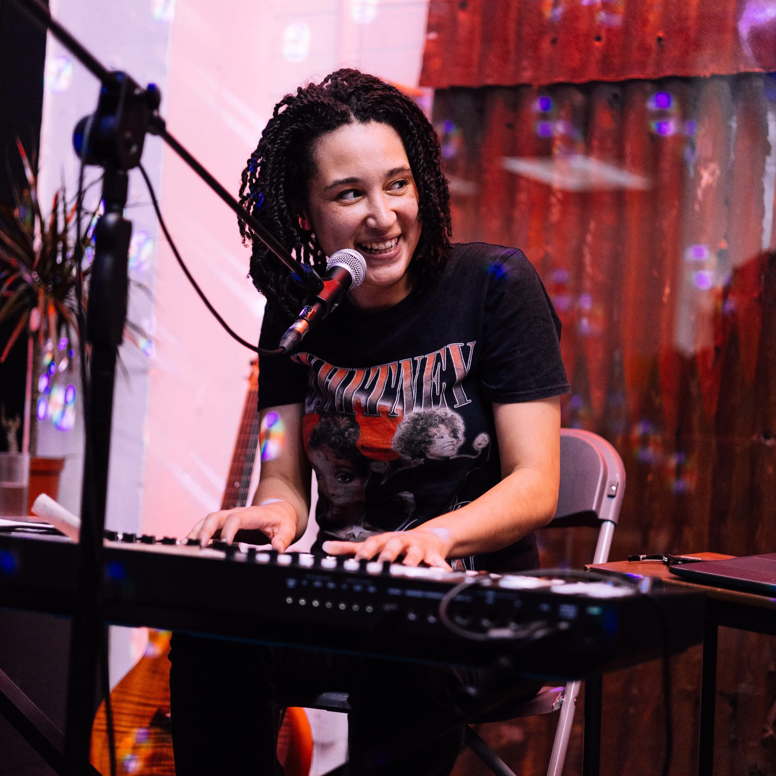 A Photo of a woman in her 30's sat playing a keyboard and smiling with her head turned to the right with a microphone at her lips. She has light brown skin, is wearing a black shirt sleeved  t-shirt with a graphic on the front.