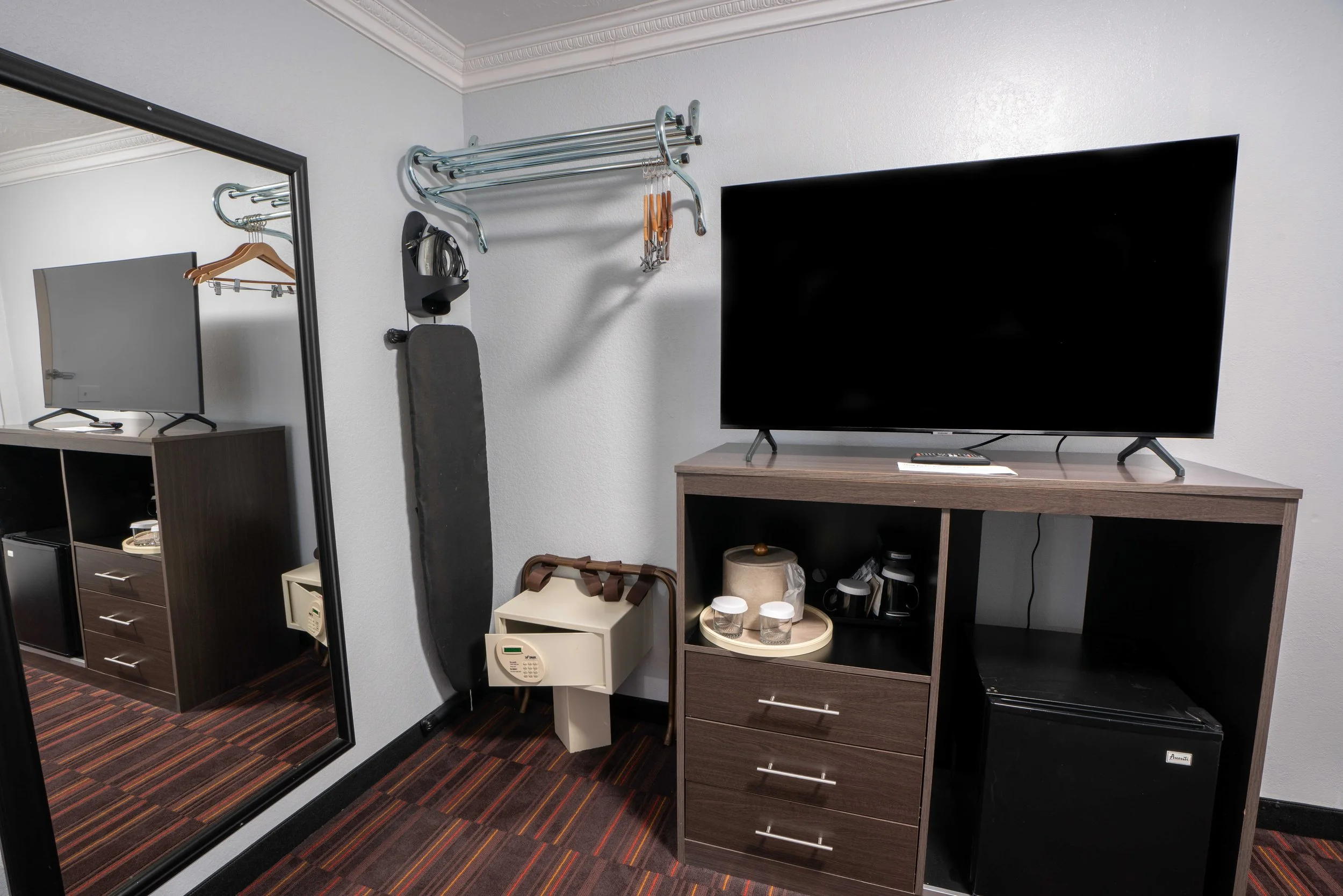 Hotel room amenities including a wall-mounted closet rack with hangers, an ironing board, a safe, a TV on a stand, a mini fridge, and a coffee setup.