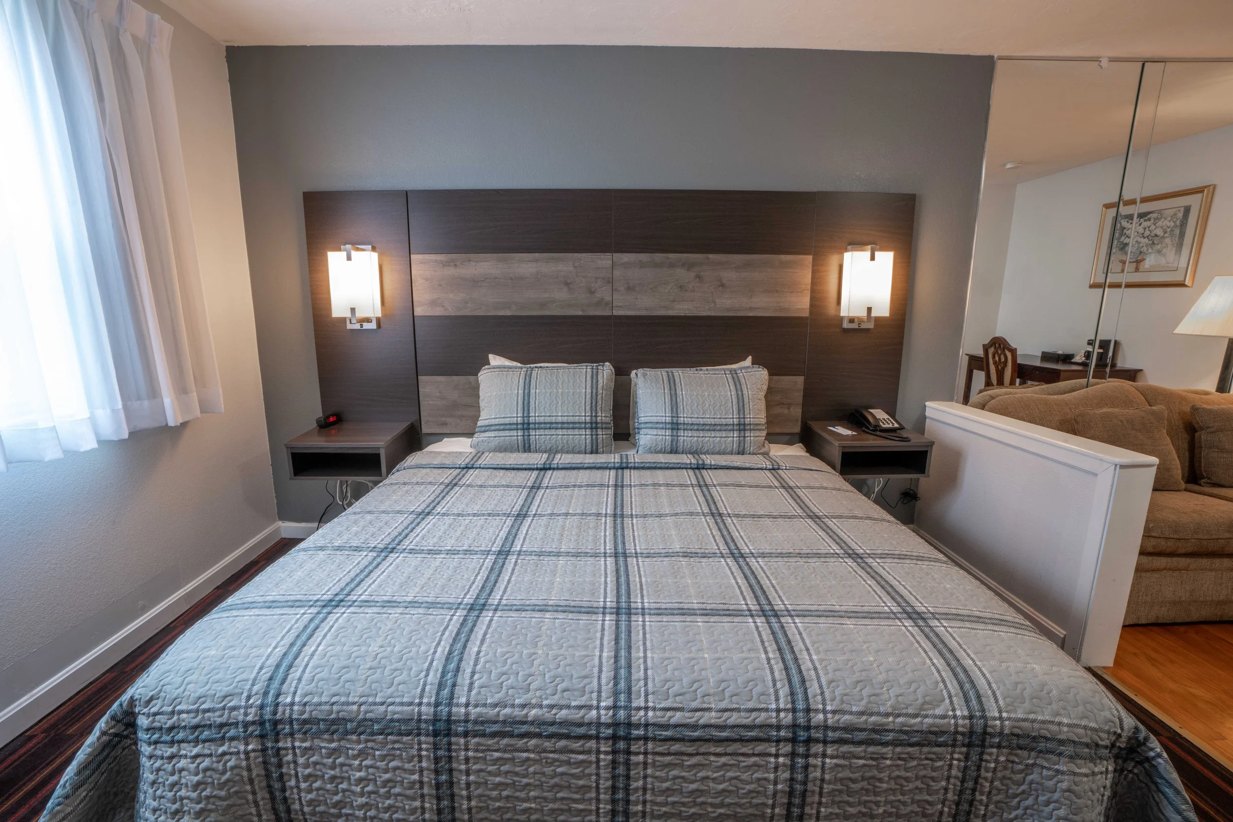Hotel room with a plaid bedspread, modern headboard, wall-mounted lamps, and bedside tables.