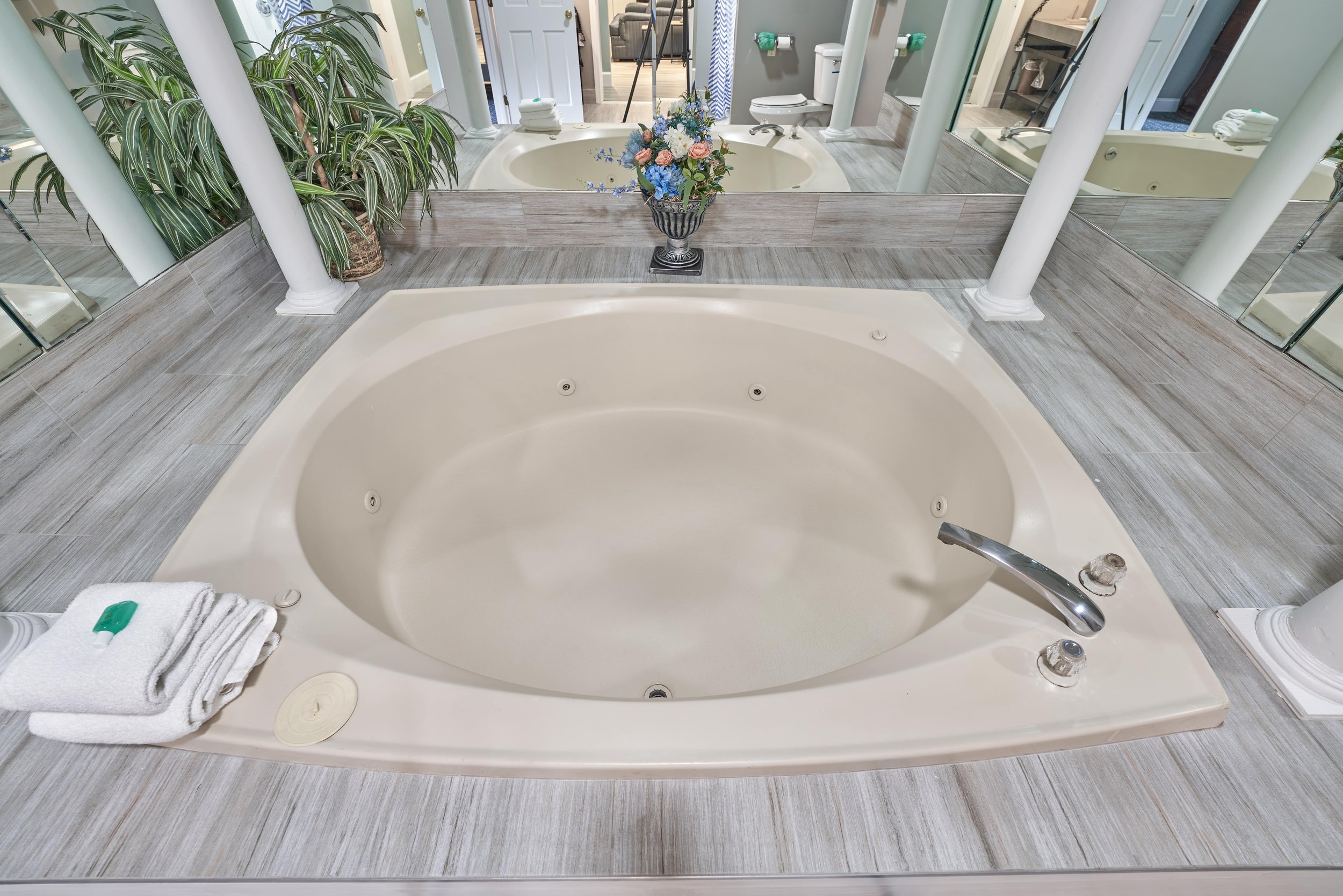 Large oval bathtub with jets, surrounded by mirrors and columns, with towels and a plant nearby.