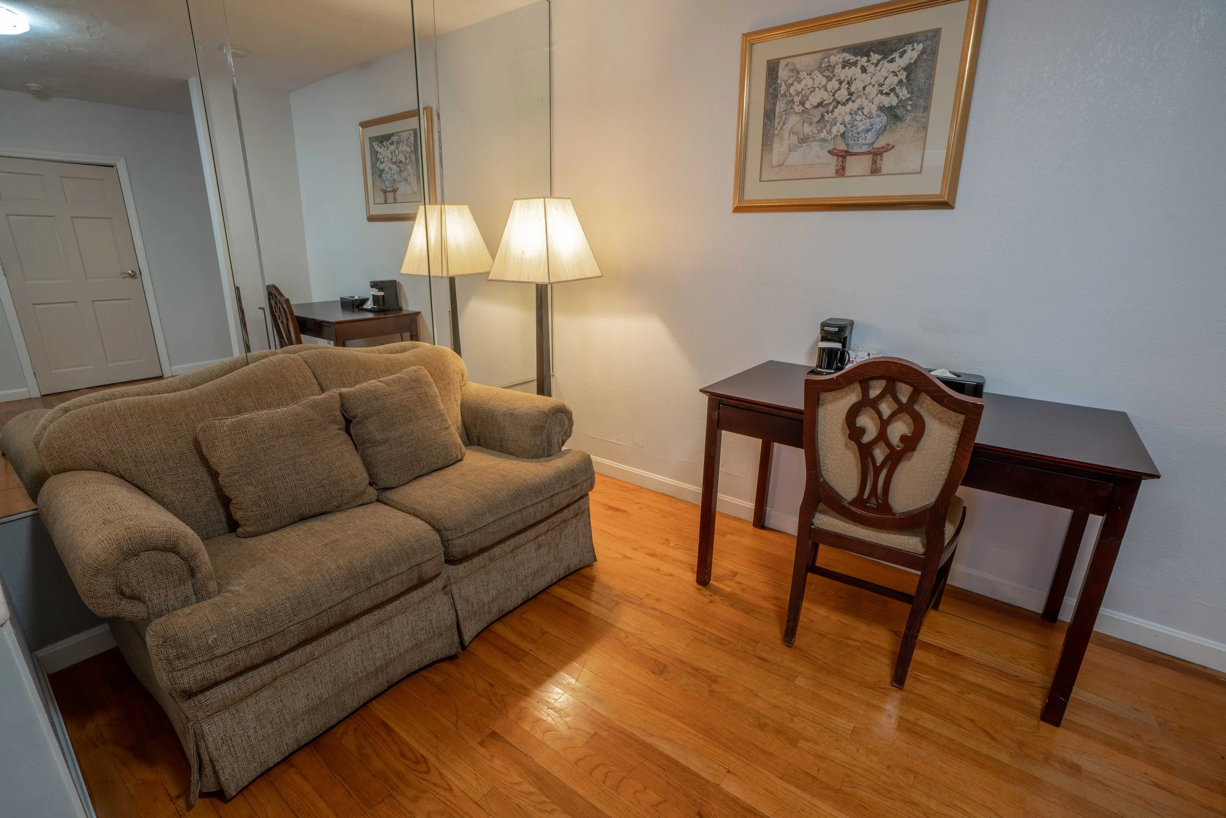 Room with a beige sofa, wooden desk, and chair on hardwood floor; a lamp and artwork are visible.