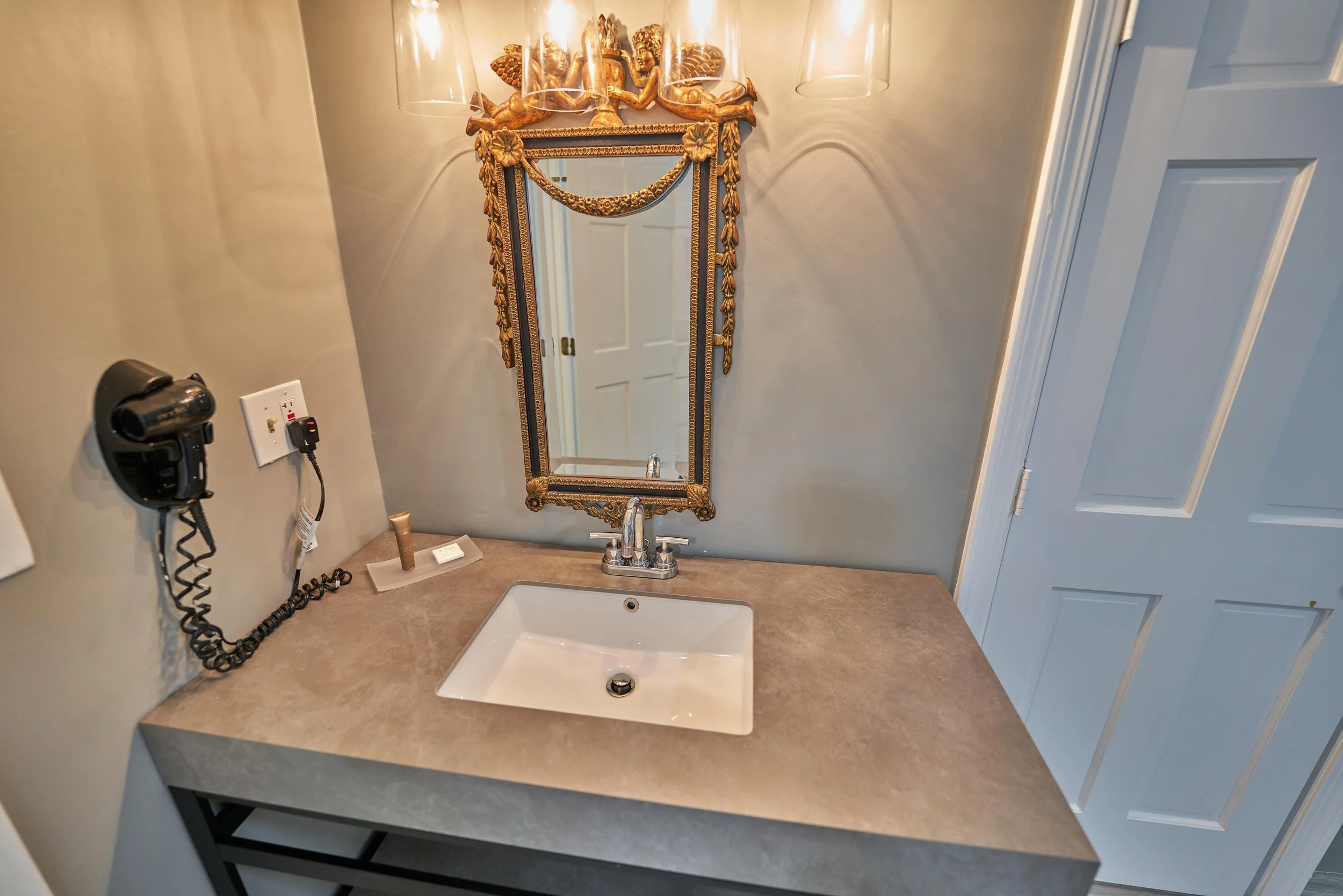 Bathroom vanity with a rectangular sink, ornate gold-framed mirror, wall-mounted hair dryer, and overhead lighting.