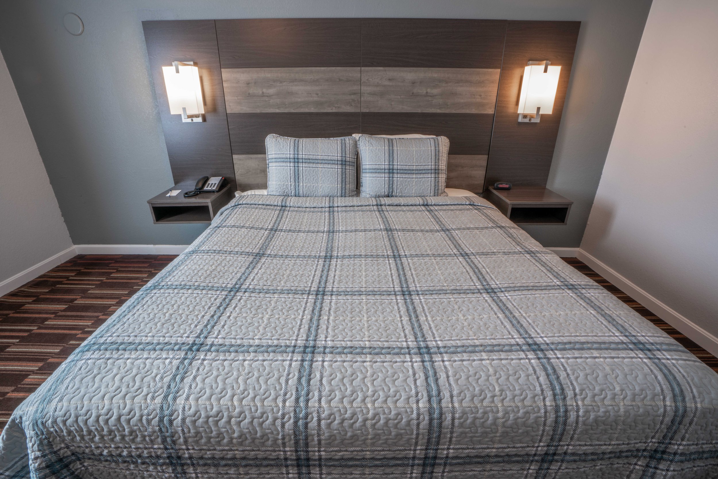 Hotel room with a bed, plaid bedding, two pillows, modern headboard, wall-mounted lights, and bedside tables.