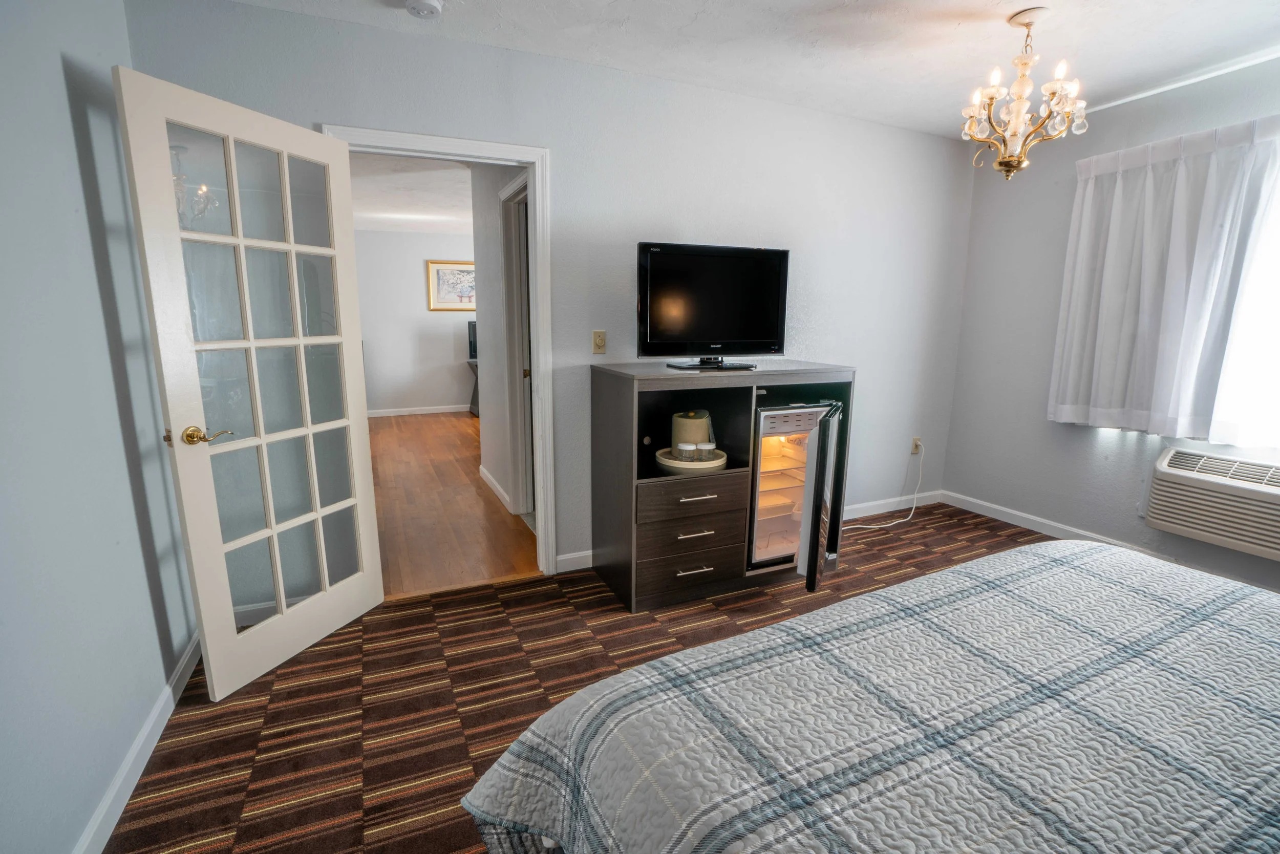 Hotel room interior with a bed, TV, mini-fridge, chandelier, and a French door.