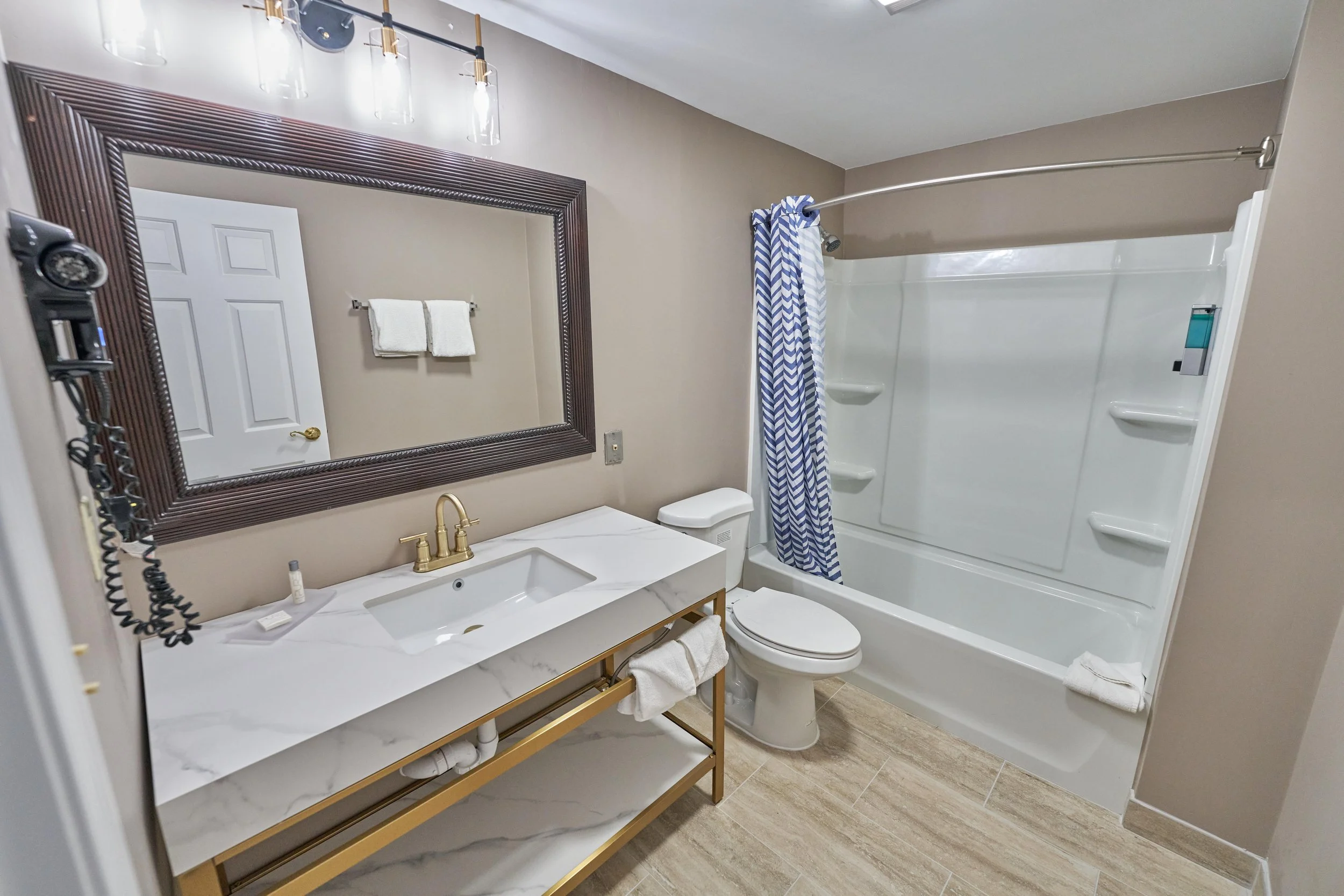 Modern bathroom with large mirror, white vanity, gold faucet, and bathtub with blue and white shower curtain.