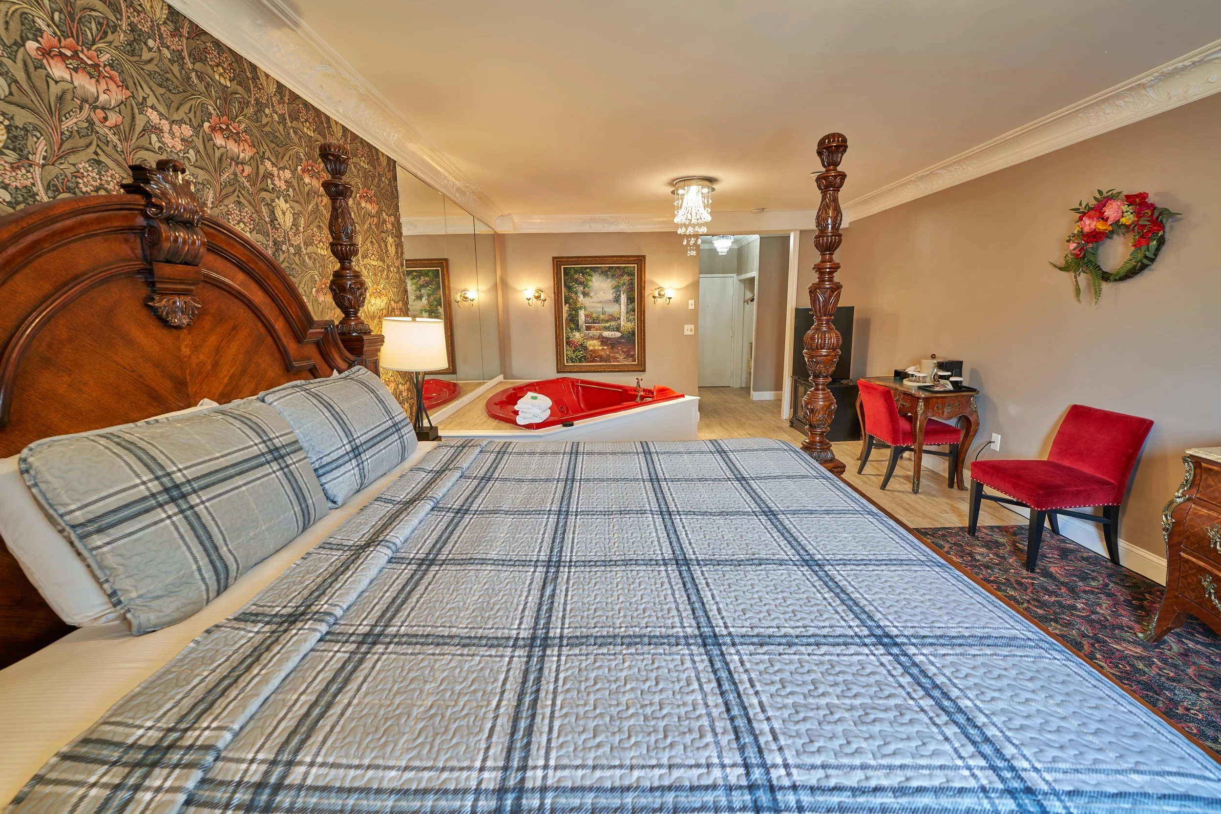 Luxurious hotel room with a large four-poster bed, floral wallpaper, red chairs, a red heart-shaped jacuzzi, and a small table with a coffee maker.