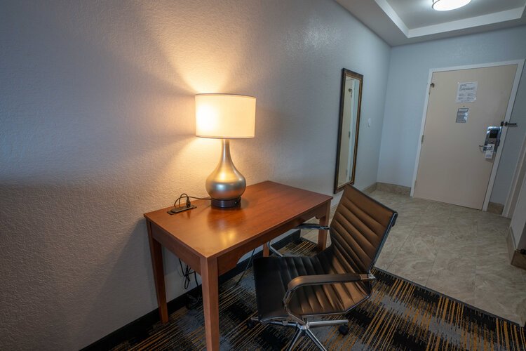 Hotel room interior with desk, lamp, chair, and mirror.