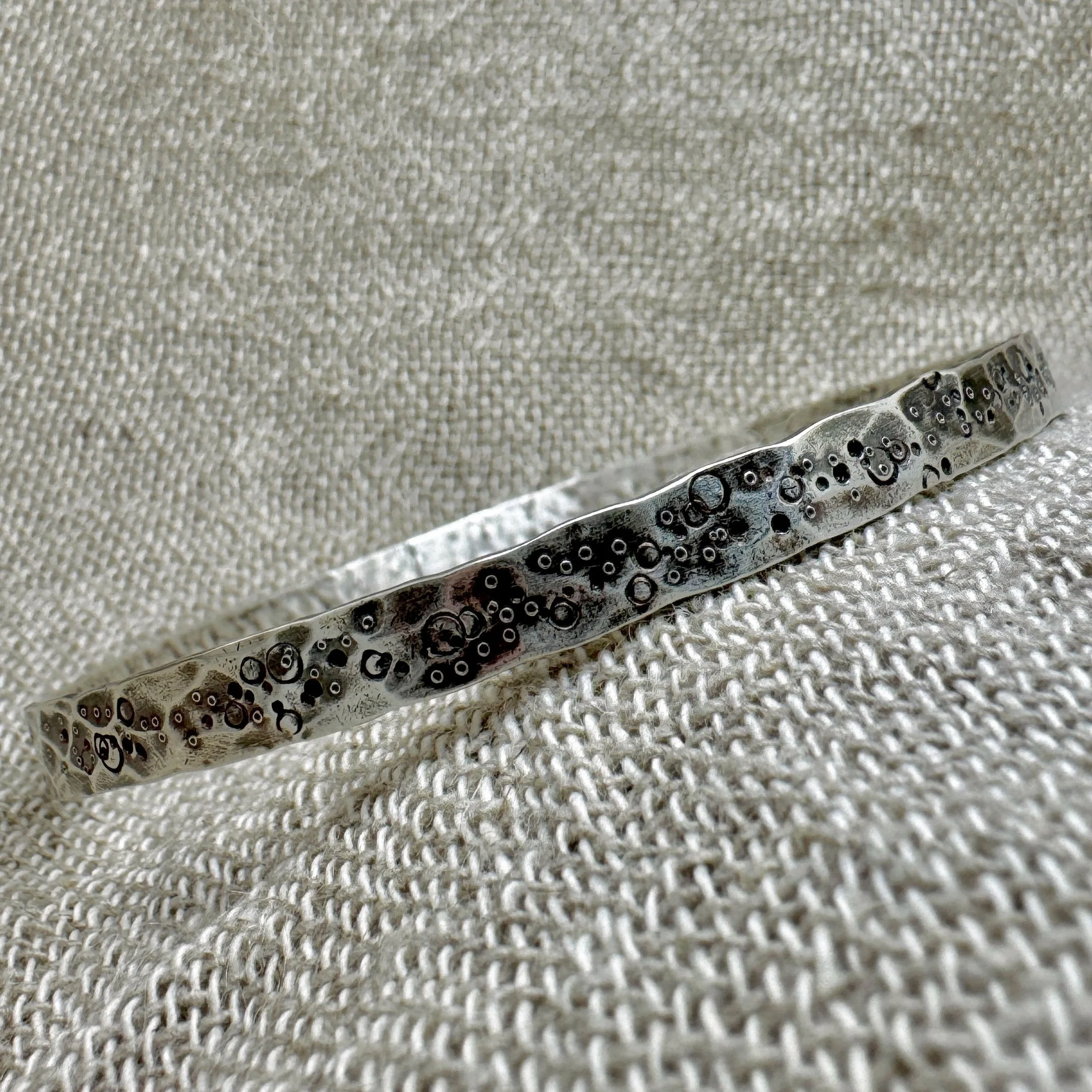 Wide textured oval silver bangle with pitted and punched markings laying on a textured fabric