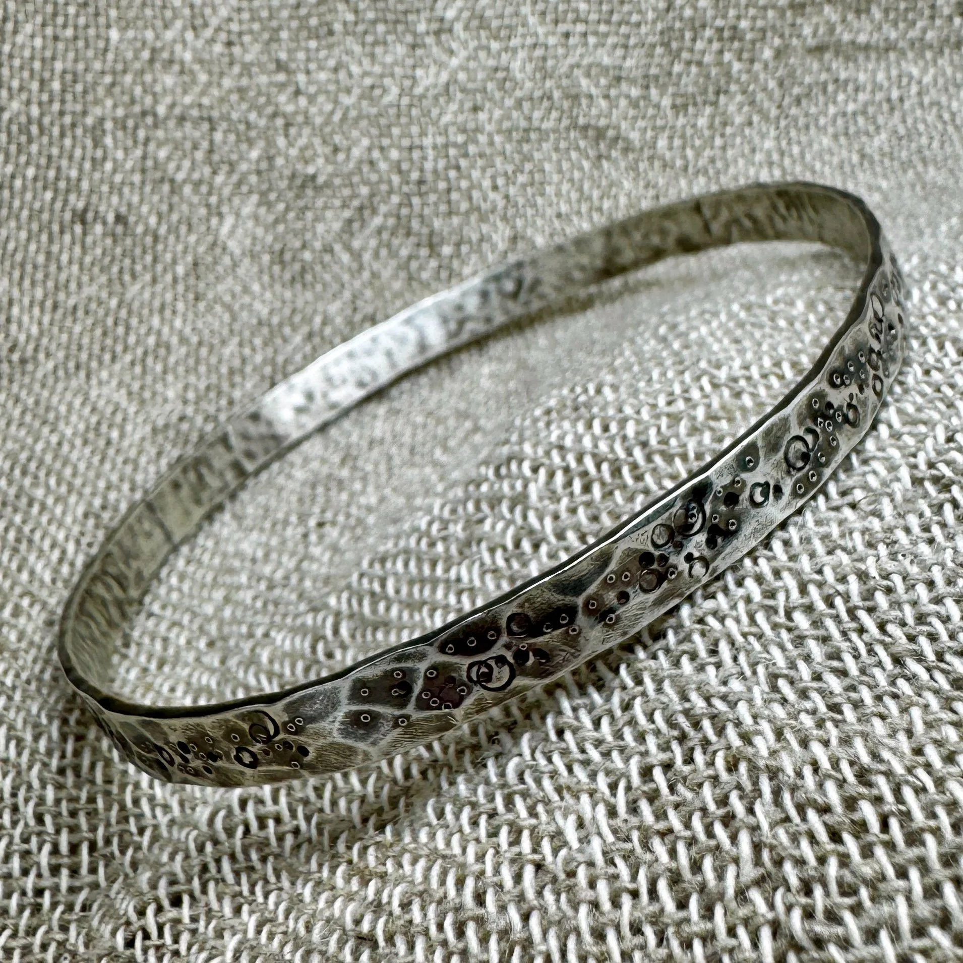Side view of a wide textured silver bangle with pitted and punched markings laying on a textured fabric background
