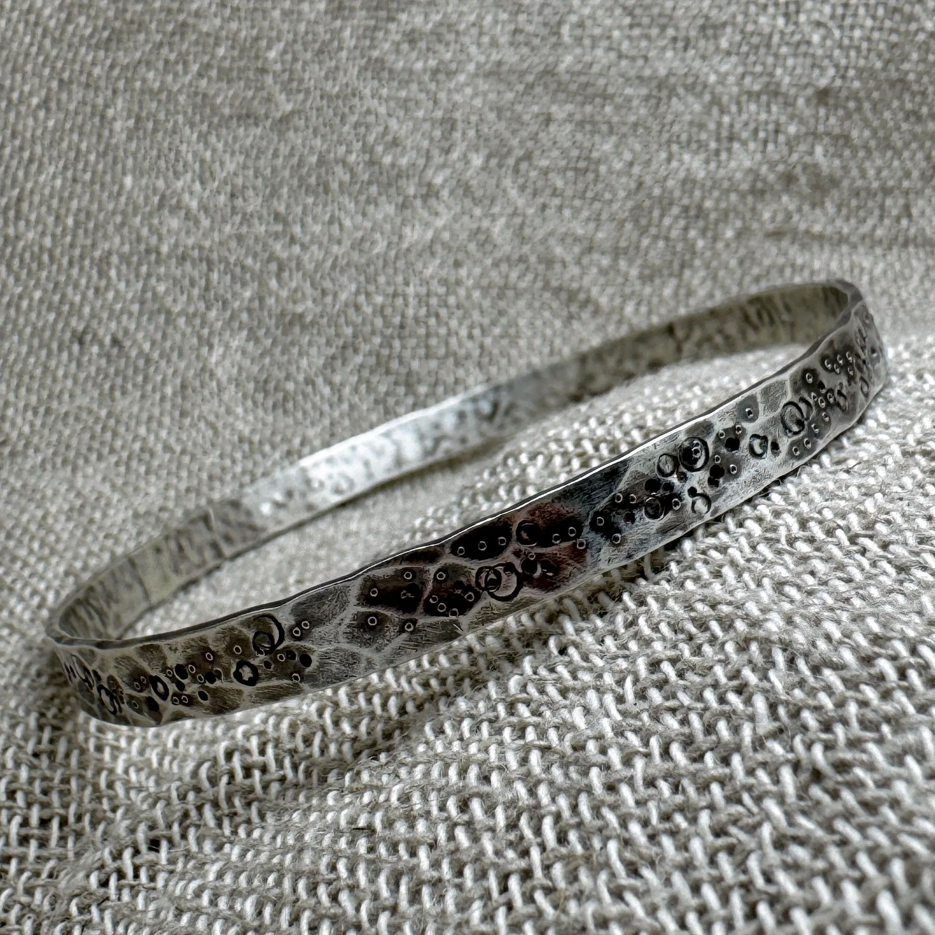Wide textured silver bangle with pitted and punched markings laying diagonally on a textured fabric