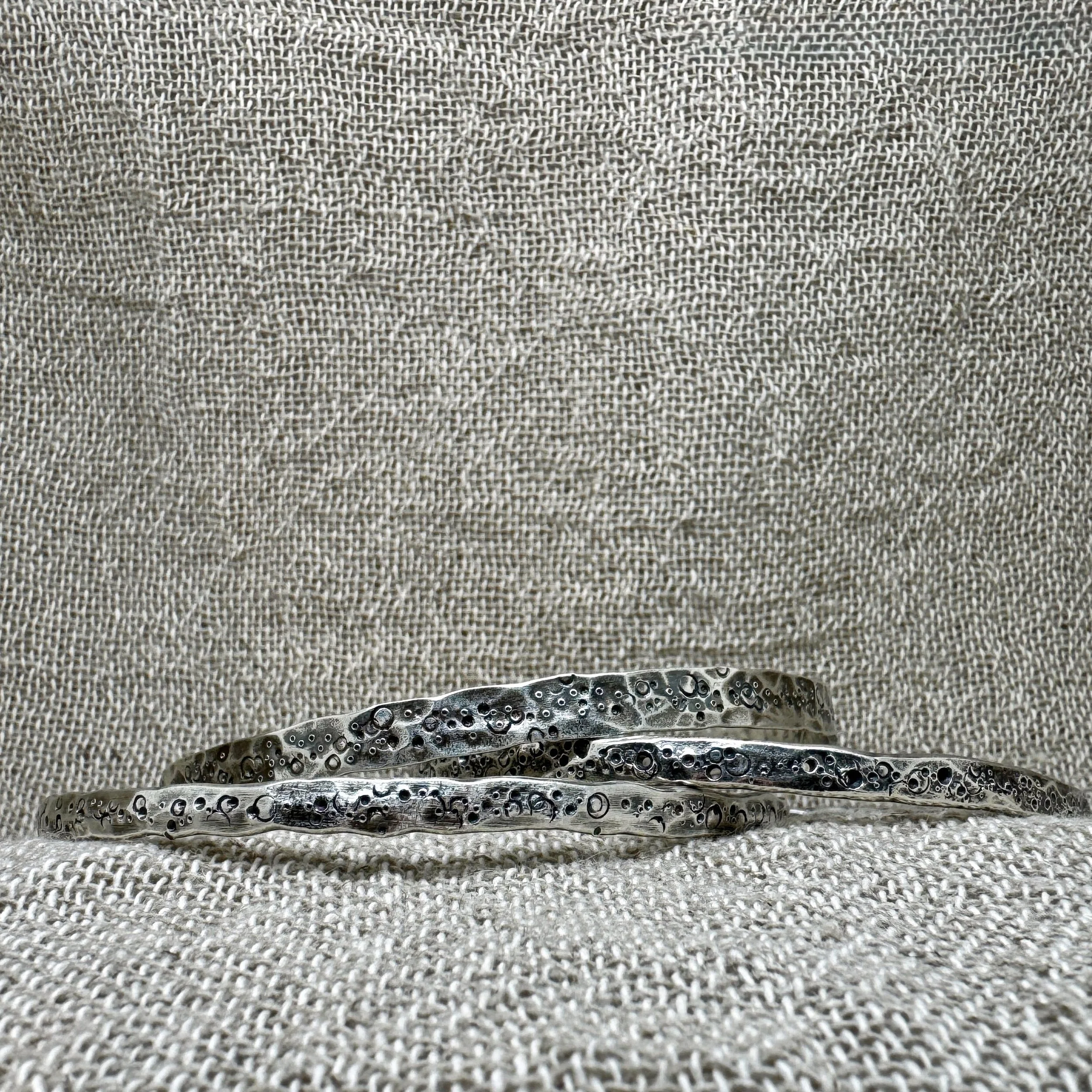 Set of three textured silver bangle with pitted and punched markings stacked up on a textured fabric