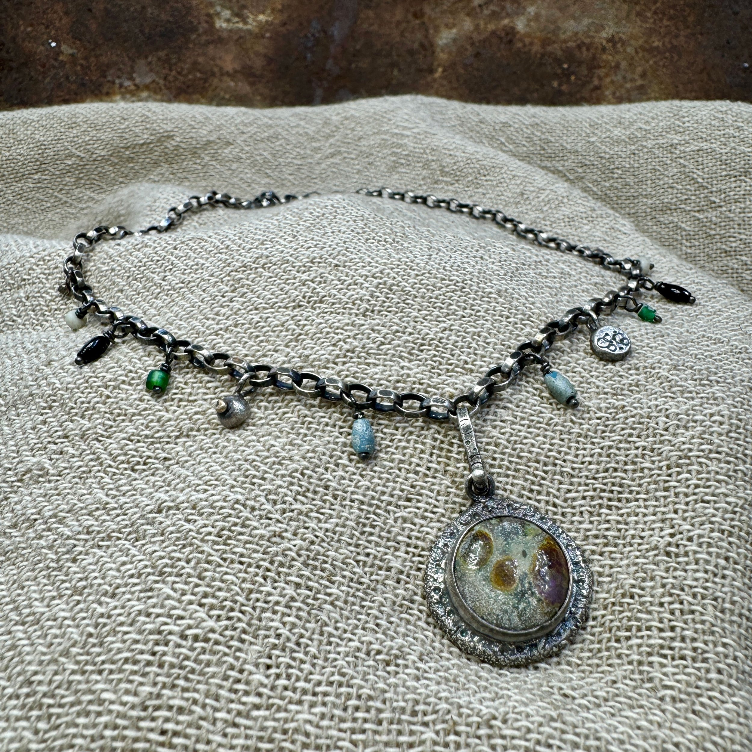 Glass pendant with rust, ochre and aqua green colours. The dark silver textured surround and chunky chain are embellished with small beads and silver charms.