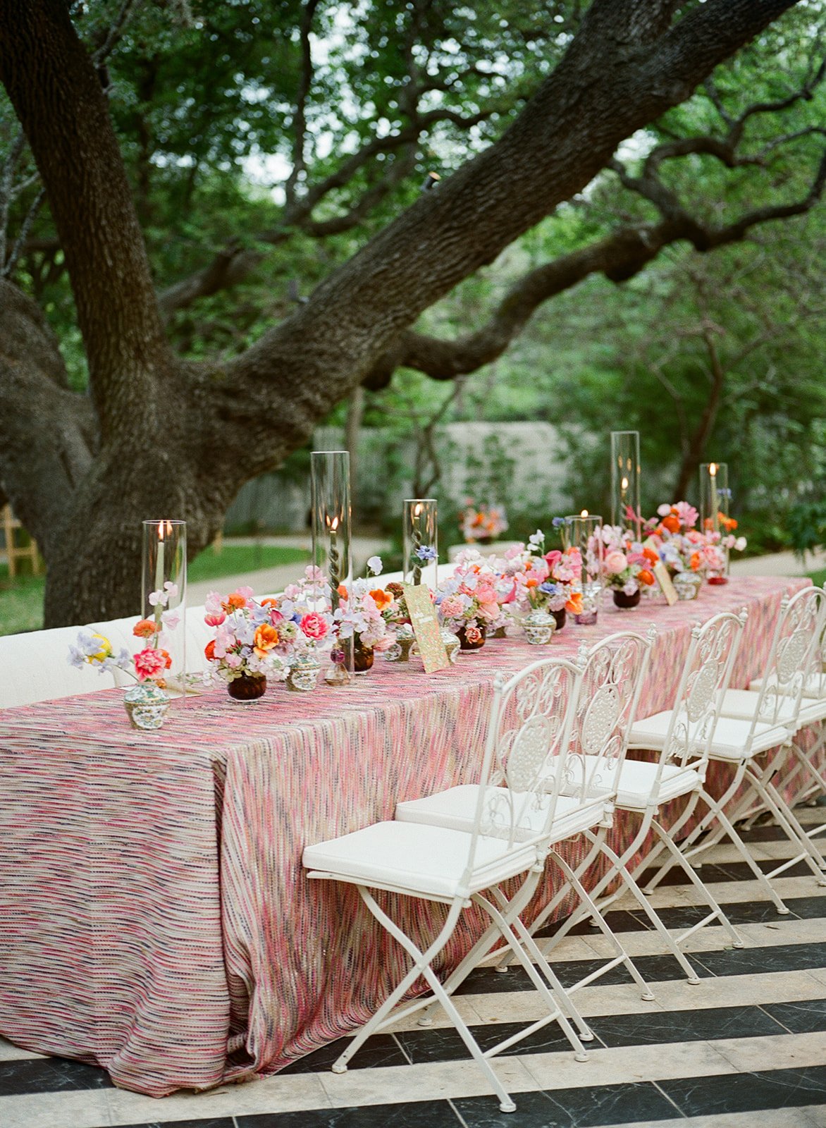  Long reception table with a striped pink tablecloth and a row of floral centerpieces under an oak tree at Hotel St. Cecilia 