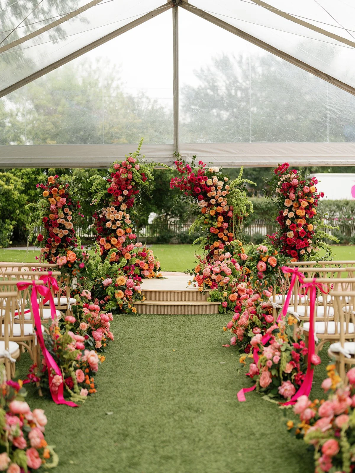  Wide view of the ceremony aisle under a clear-top tent, with a red, coral, and pink floral arch at the end and pink ribbon streamers tied to the cane chairs at Woodbine Mansion 