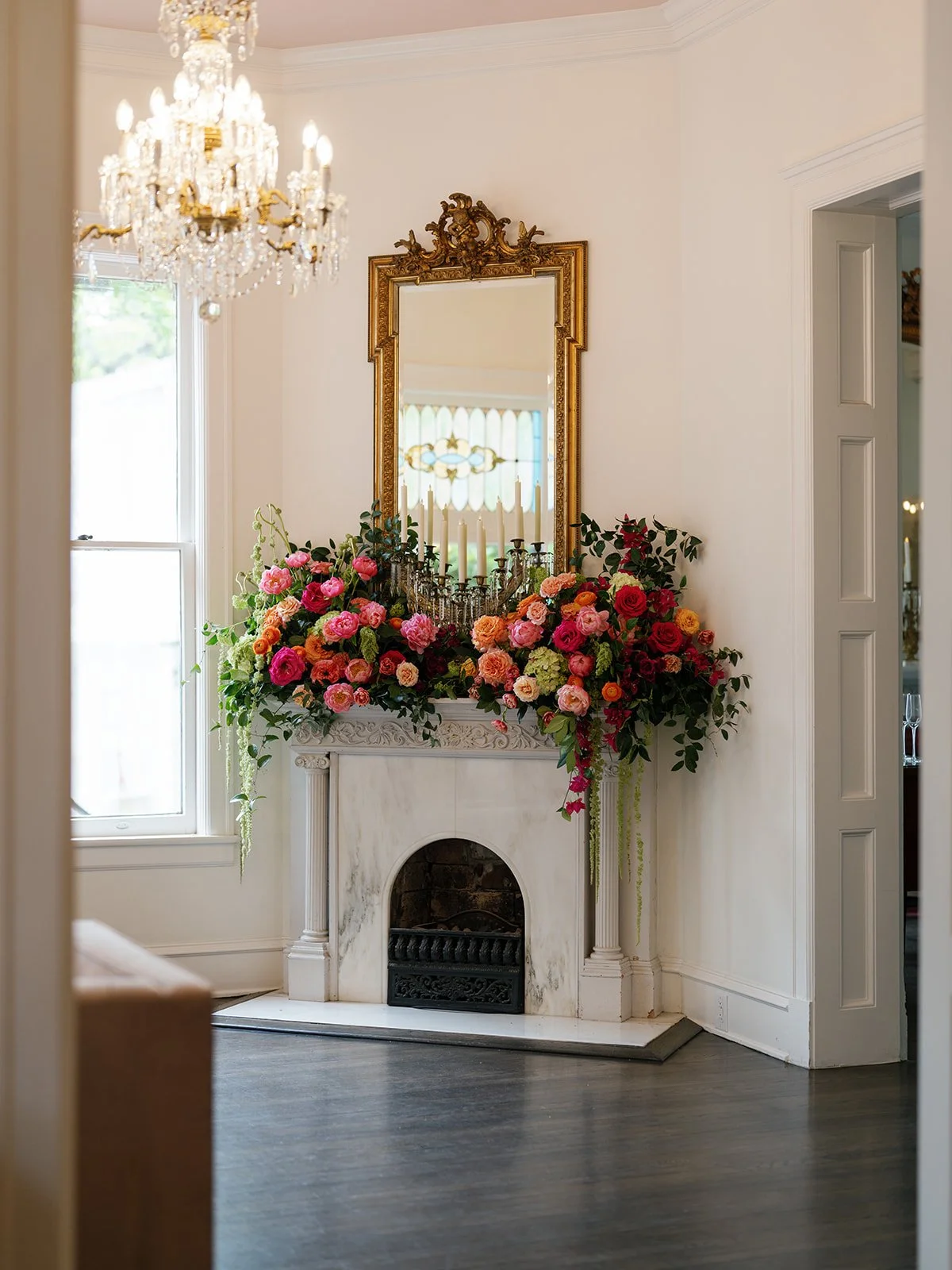  Marble fireplace mantel with a cascading arrangement of red, pink, and orange roses beneath a gold mirror and crystal chandelier at Woodbine Mansion 