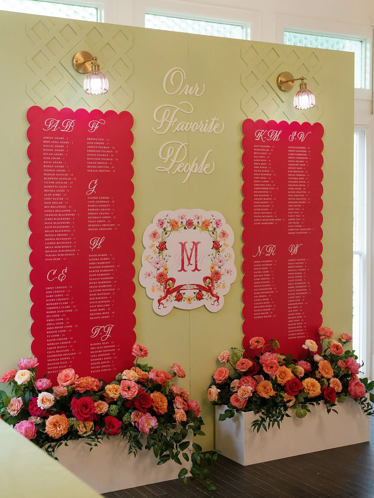  Personalized seating chart display with alphabetical guest cards and a floral-wreathed monogram, flanked by coral and red rose planters at Woodbine Mansion 