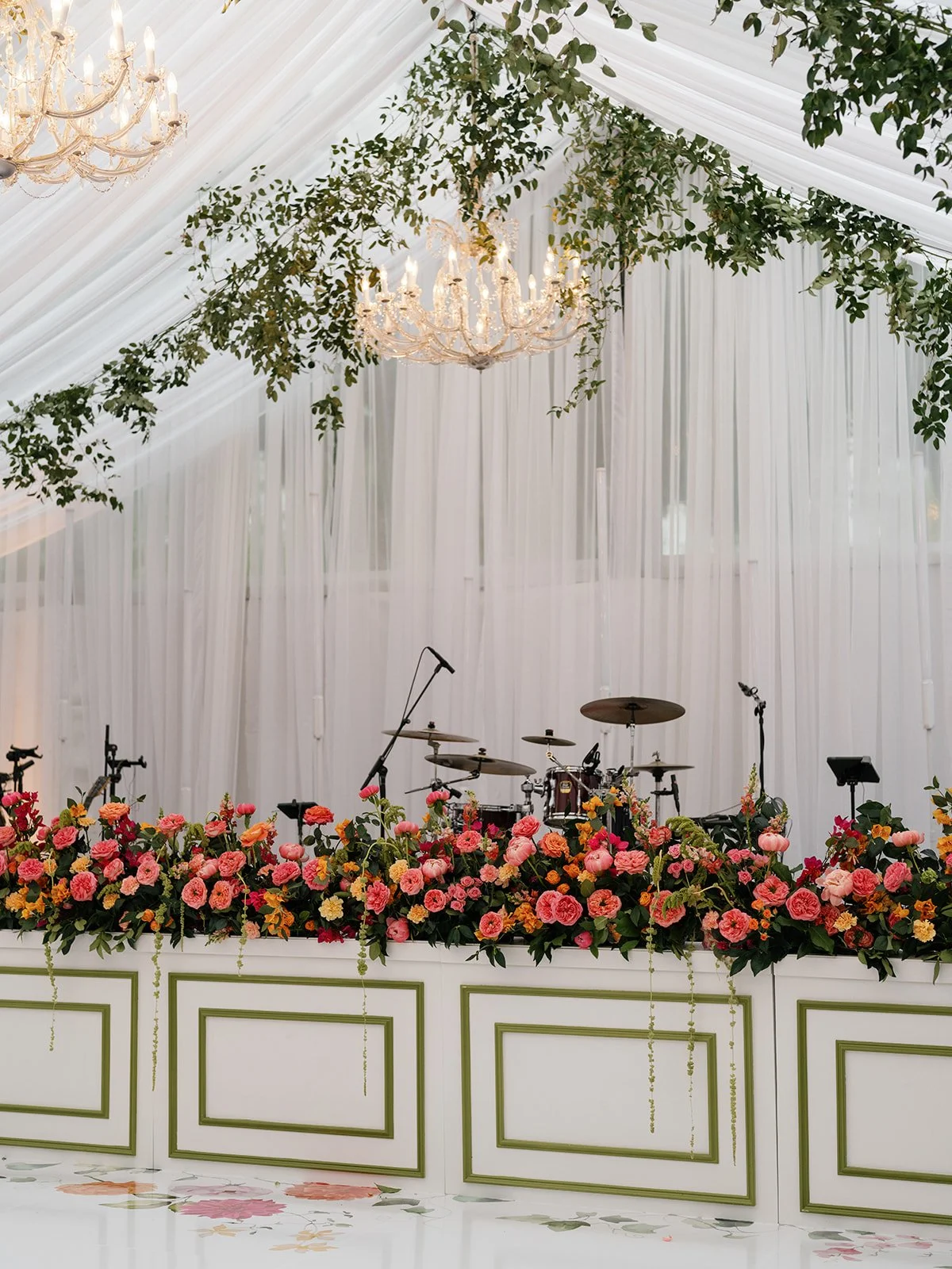  View toward the band stage under the tent, with trailing smilax overhead and a long coral and red floral runner across the stage front at Woodbine Mansion 