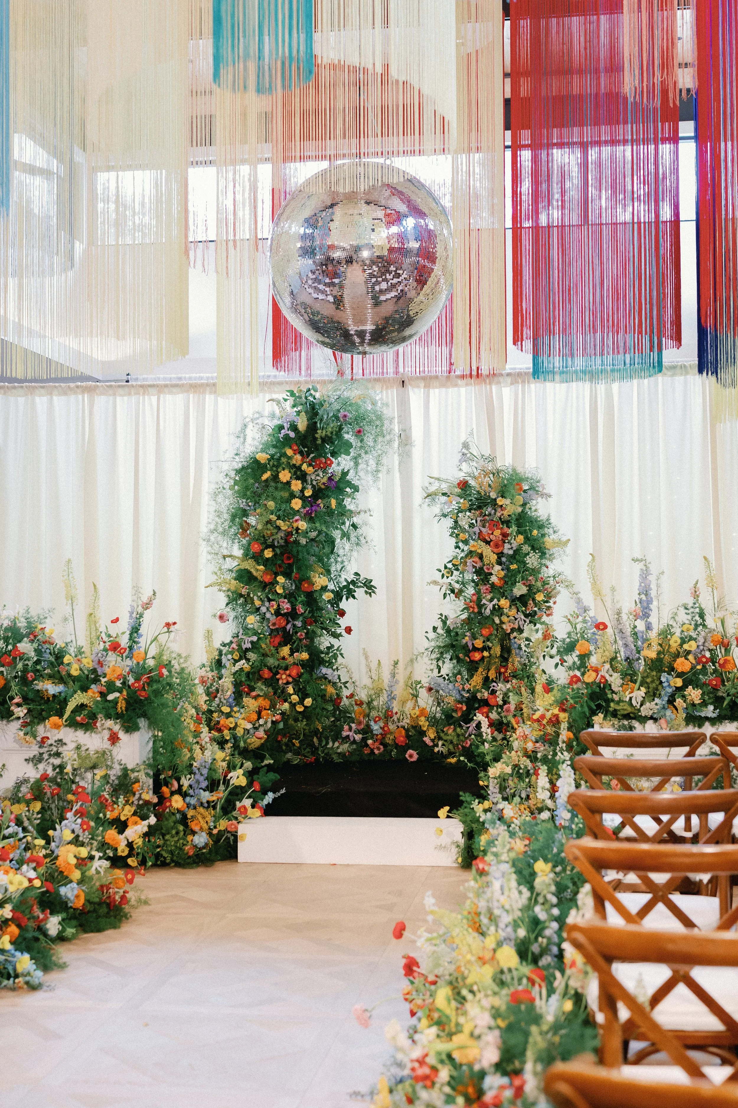  Wide view of the ceremony aisle at Mattie's with a colorful garden-style floral runner leading toward two tall floral columns and a disco ball above 