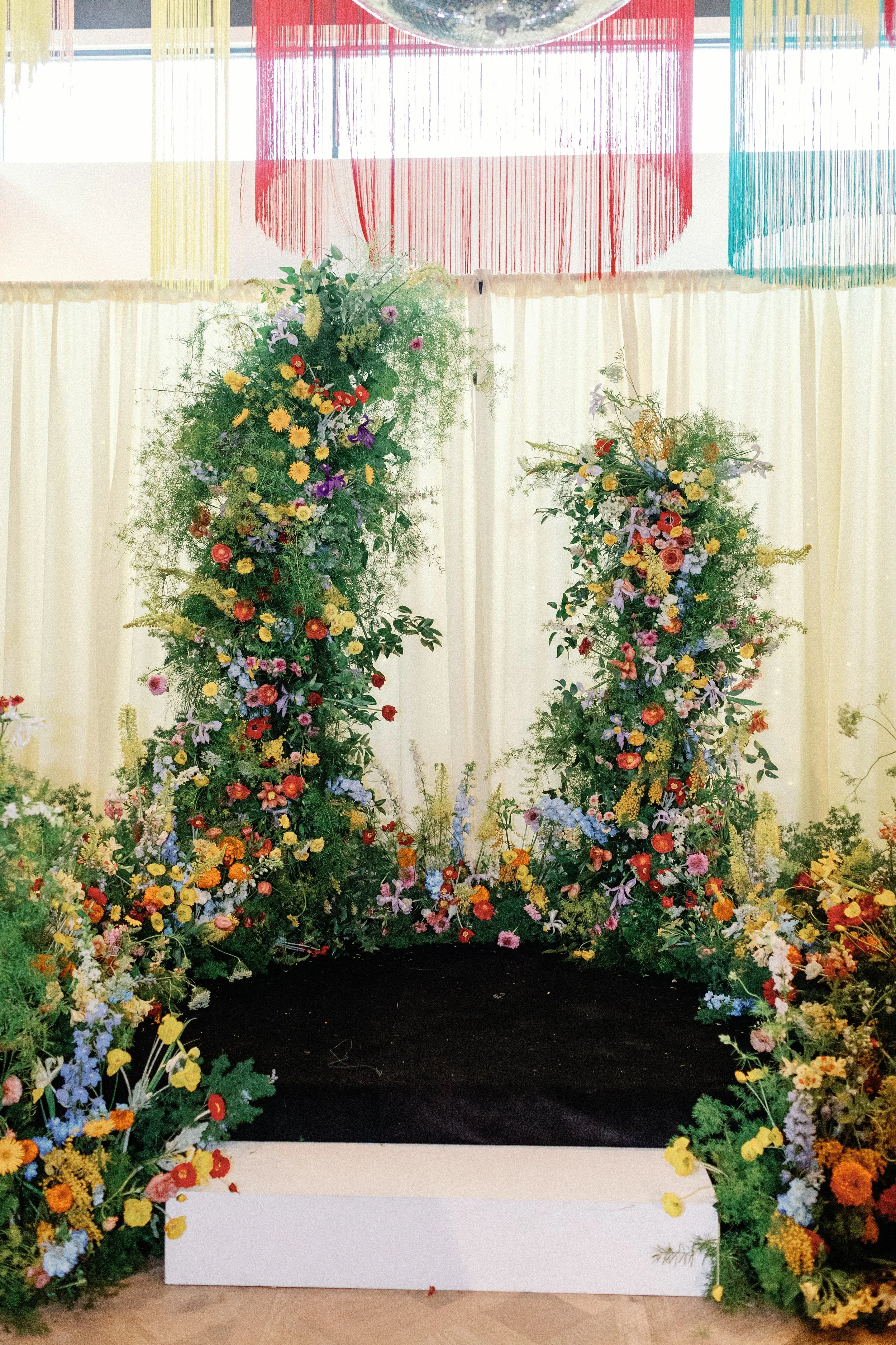 Two tall asymmetrical floral columns framing the altar area at Mattie's, with a rainbow fringe backdrop behind them 
