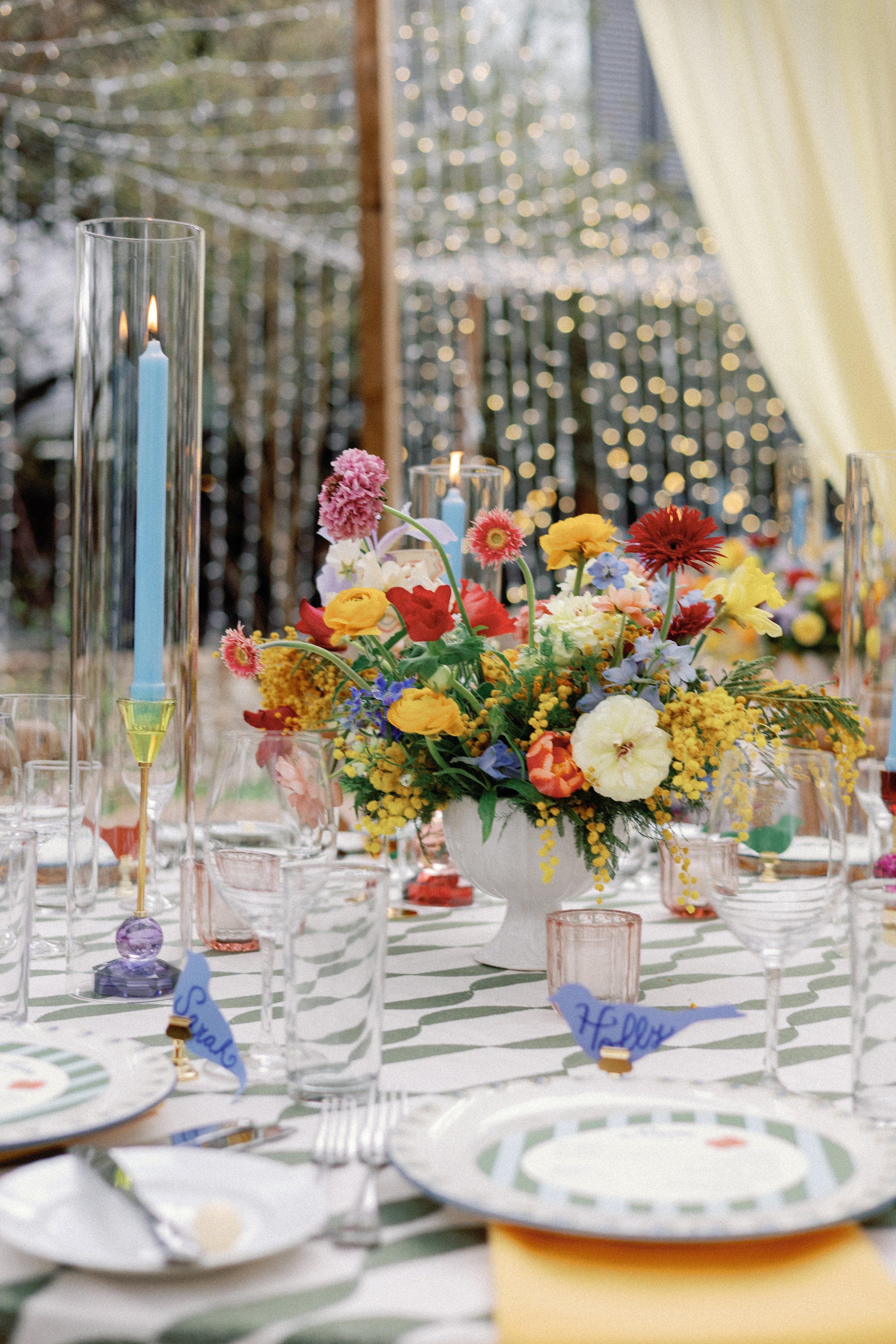  Table setting with a vibrant yellow, red, and purple floral centerpiece, striped plates, and a blue taper candle at Mattie's 