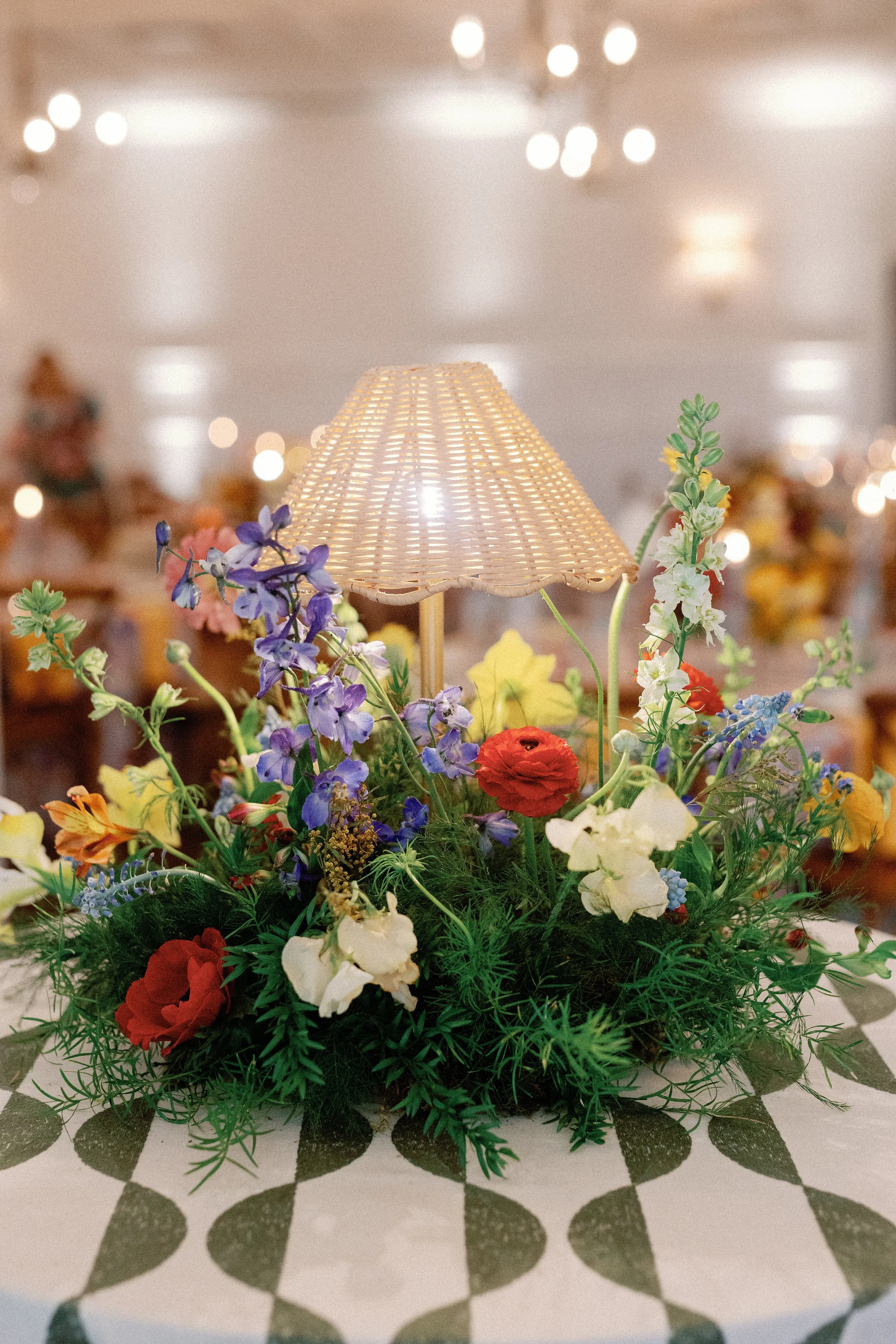  Close-up centerpiece with a wicker lamp shade and purple, red, and yellow blooms on a checkered green and white tablecloth at Mattie's 