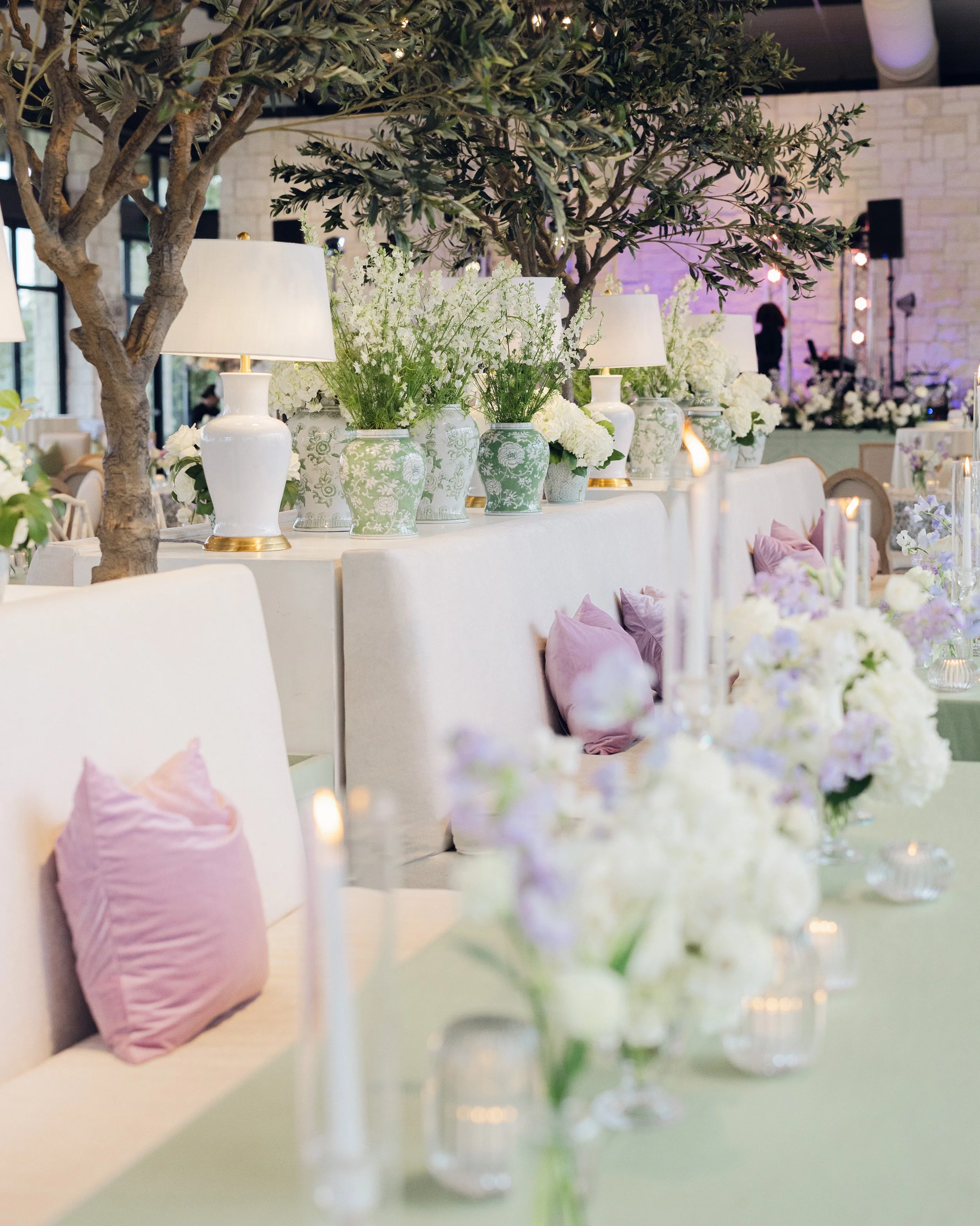  Reception table with faux olive trees, white ceramic lamp style vases, and lavender pillows against white banquette seating 