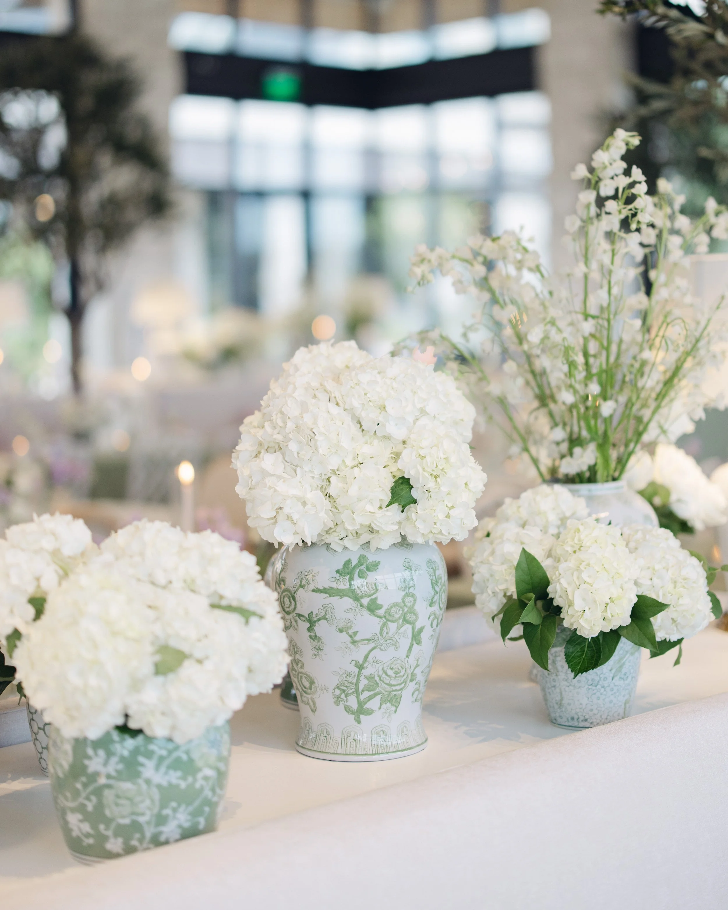  Cluster of white ceramic vases and ginger jars filled with white hydrangea on a patterned linen tablecloth 
