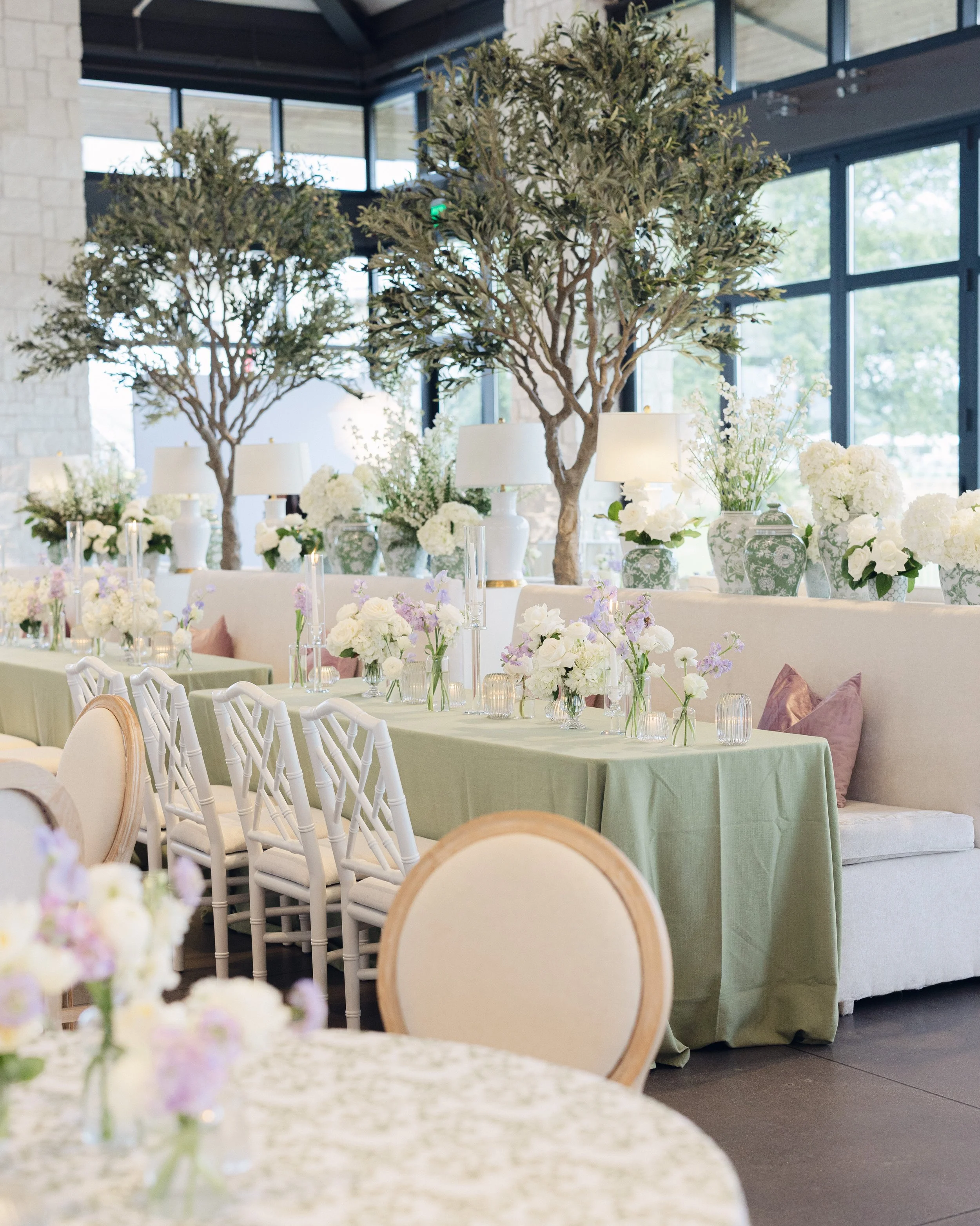  Wide shot of Omni Barton Creek reception tables with sage green linens, lavender pillows, and olive trees under floor to ceiling windows 