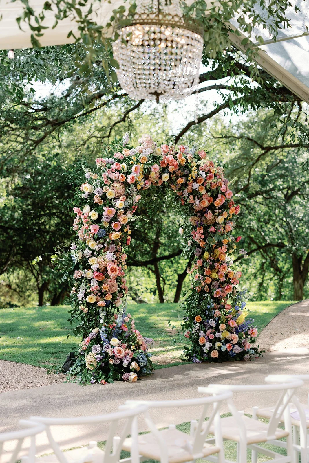  Floral ceremony arch in coral, pink, and yellow hanging beneath oak trees with a crystal chandelier above 