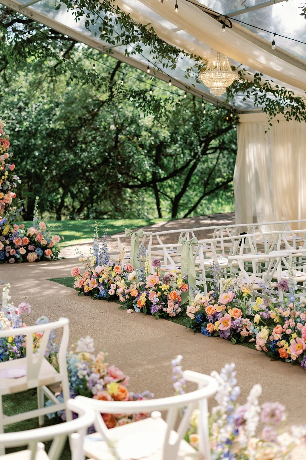  Tented wedding ceremony aisle lined with white chairs and colorful floral arrangements, oak trees visible through the tent 