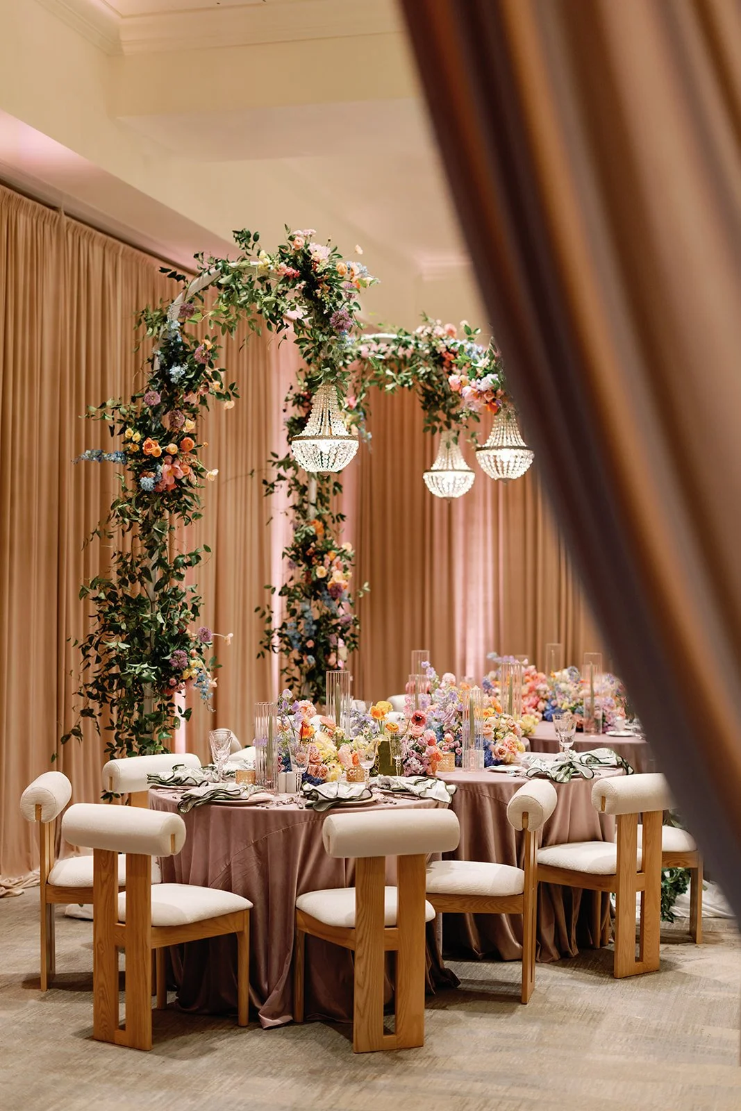  Long reception table with wood and cream chairs beneath hanging floral installations and crystal chandeliers 