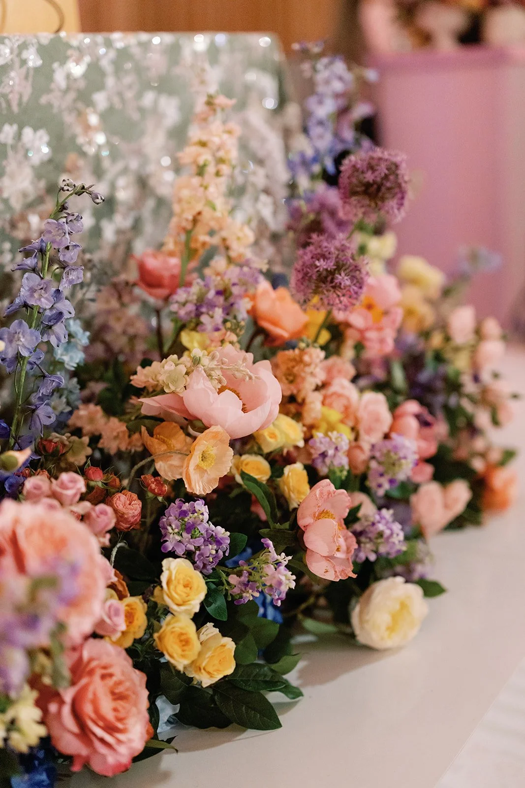  Close up floral arrangement in peach, lavender, and blue tones on a light pink surface 