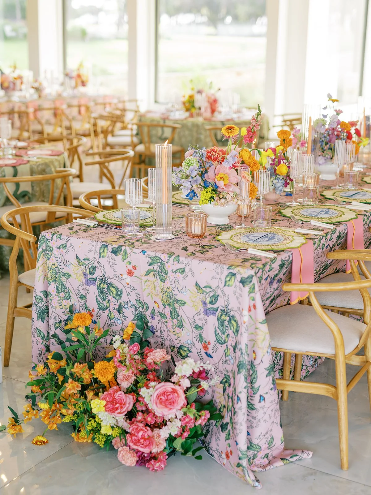  Long reception table with floral print linen, wood chairs, and a colorful centerpiece spilling onto the floor 